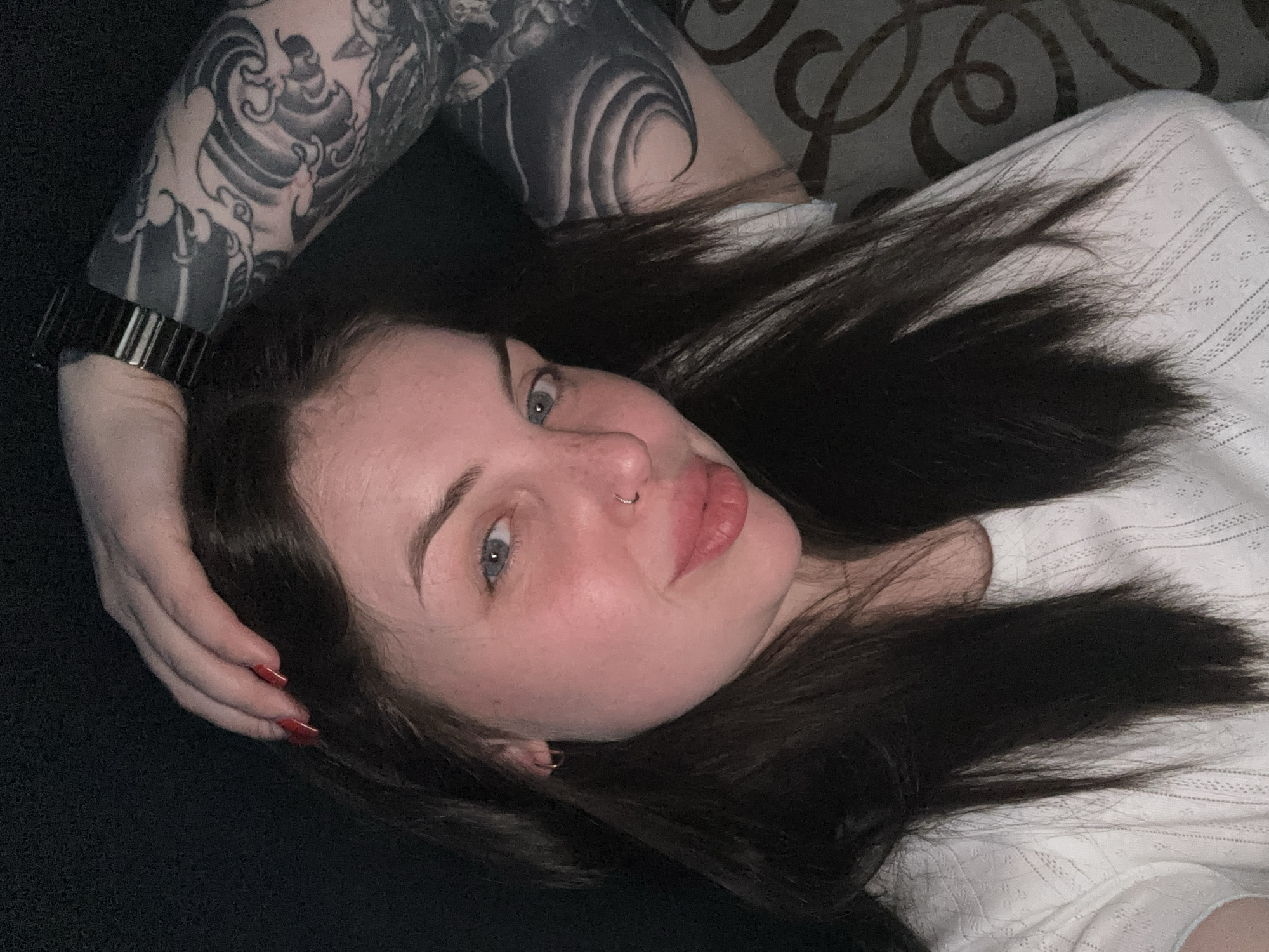 A person is lying down, angled slightly towards the viewer. They have dark hair, fair skin, and a nose ring. The arm closest to the viewer has a tattoo covering a significant portion of it. A black watch band is visible on their wrist. They are wearing a white, textured shirt. The background appears to be a mix of dark solid and patterned surfaces. There is no information to indicate the picture was taken in Chornomorskoe, Ukraine.
