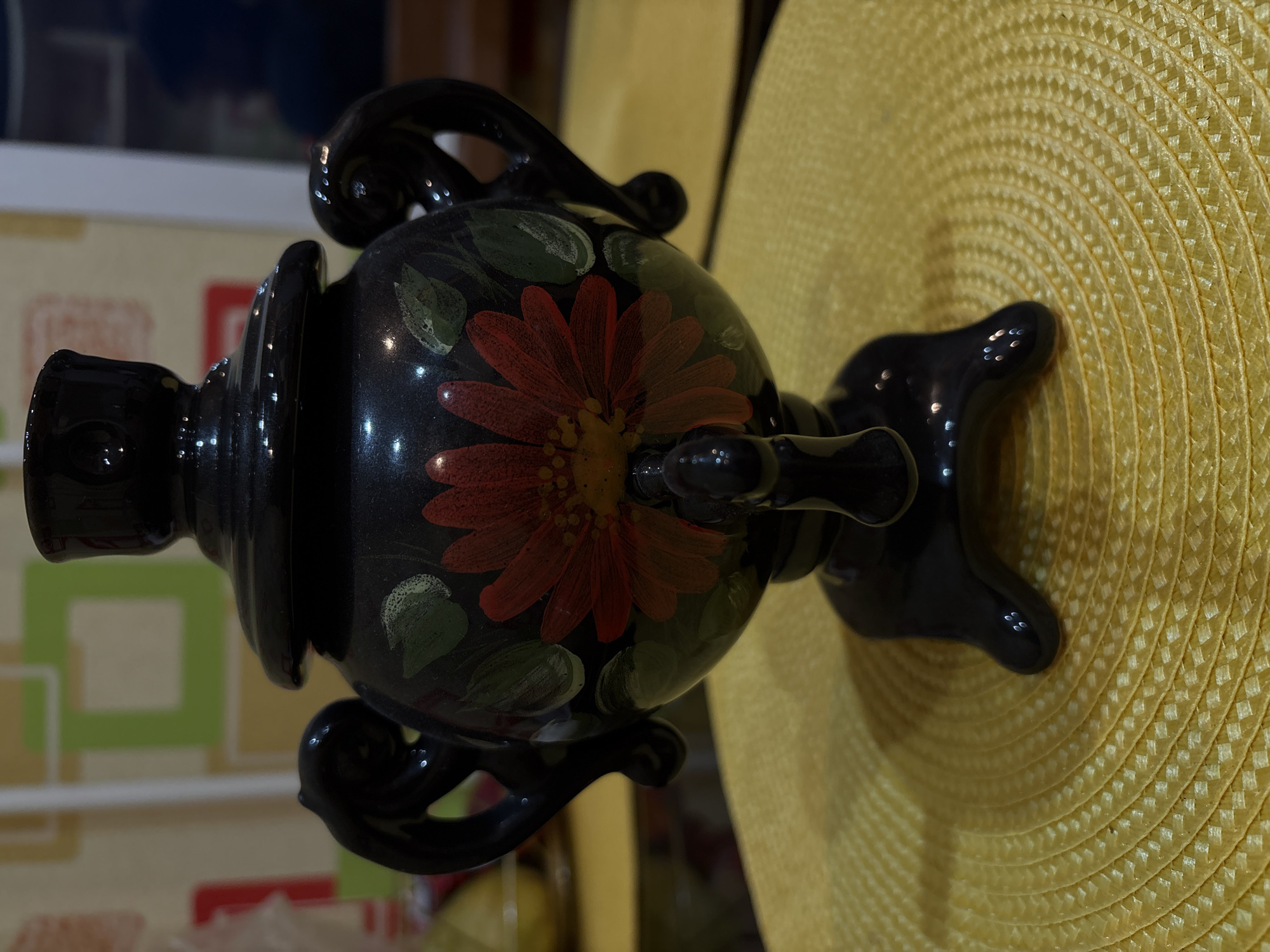 A black samovar with a red and yellow flower painted on it sits on a yellow placemat. The samovar has two black handles and a spout. The background is blurry and appears to be a wall with a colorful geometric pattern. The lighting is dim, and there are reflections on the surface of the samovar. The image is taken from a slightly elevated angle, looking down at the samovar. There are no people or actions depicted in the image. There is no information available about the specific location in Vitsebsk, Belarus.
