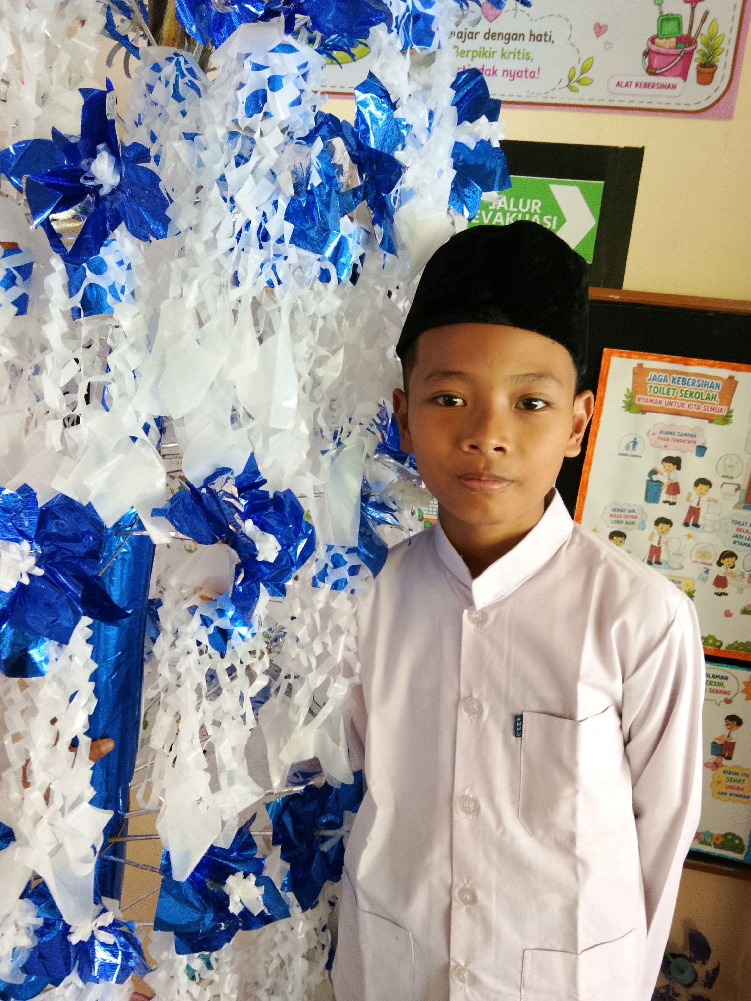 In Besusu Timur, Indonesia, a young boy stands in front of a wall decorated with blue and white streamers. He wears a white long-sleeved shirt with a black peci hat. Behind him, a bulletin board displays information about school sanitation, with illustrations of children participating in cleaning activities. The overall environment appears to be an educational institution, possibly a school hallway or classroom, given the presence of the bulletin board and the decorative streamers which might be part of a celebration or event.