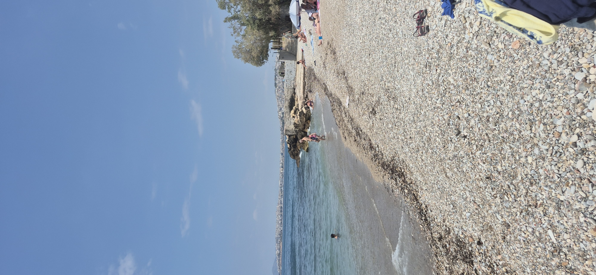 The image depicts a scenic view of a pebble beach in Paleo Faliro, Greece, with the city skyline visible in the distance across the shimmering blue sea. The sky is a clear, bright blue with a few wispy clouds. On the beach, several people are enjoying the sunny day. In the foreground, the pebble-strewn shore is evident, with a few pieces of dark seaweed scattered along the waterline.
Further down the beach, a group of individuals are relaxing. Some are lying on towels, sunbathing, while others are seated, enjoying the atmosphere. A white umbrella provides shade for some of the beachgoers. Closer to the water, a man and a child are wading in the shallows. To the left, a young boy is swimming in the sea. On the right side of the frame, a pair of sandals and some folded clothing are placed on the pebbles. The natural rocky formations and vegetation add to the picturesque coastal landscape.