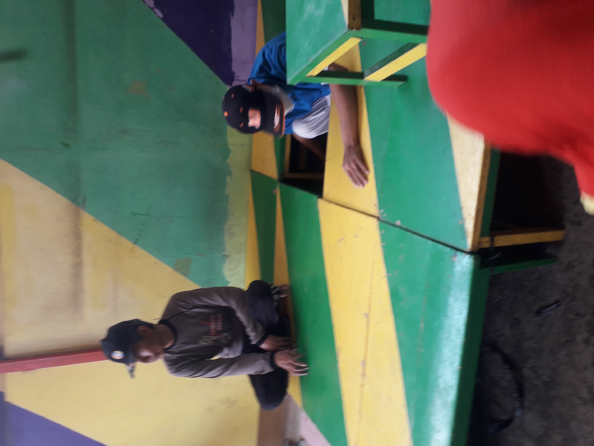The image depicts an indoor scene with two individuals, likely children or young adults, interacting in a playful or learning environment. The setting appears to be a recreational area with brightly colored, multi-level platforms and walls. The dominant colors are yellow and green on the floor, with sections of purple and yellow on the wall.
On the left, a person wearing a black cap and a gray long-sleeved shirt is sitting cross-legged on a yellow and green patterned platform. They are looking to their left, with their hands resting on their knees. The person's attire suggests casual wear.
To the right, another individual, also wearing a black cap, is partially obscured by a green and yellow platform. This person is dressed in a blue long-sleeved shirt and white pants. They appear to be reaching into a hole in the platform, possibly interacting with something or someone below. A green and yellow table-like structure is visible in the foreground on the right, with its leg extending downwards.
The floor is made of modular square panels arranged in a checkerboard pattern of yellow and green. Some of these panels have been removed or are designed to create openings. The lighting in the scene is somewhat dim, with a slight blur, suggesting it might be a photograph taken with a mobile phone in low light conditions.
The background wall is painted with large geometric shapes in yellow, green, and purple. A red beam or support structure is visible vertically on the left side of the wall.
The overall impression is that of a children's play area, an indoor playground, or a creative learning space, possibly located in Cankuang, Indonesia, given the contextual information. The activity suggests engagement and interaction, possibly a game or educational activity.