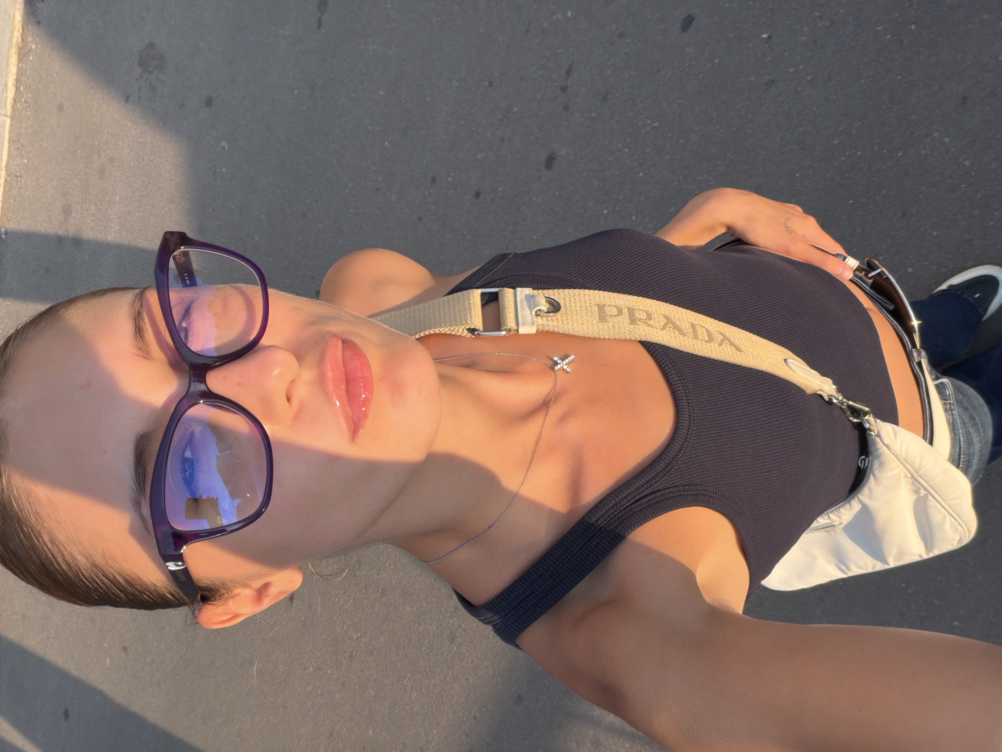 A woman stands outdoors on a paved surface, possibly a sidewalk or road, bathed in the warm glow of sunset. She is wearing a dark tank top, blue jeans, and a light-colored crossbody bag with a beige strap that has the word "PRADA" printed on it. She has on purple-rimmed glasses and a delicate silver necklace with a cross pendant. Her hair is pulled back, and she is looking slightly away from the camera. The lighting casts long shadows across the ground, indicating the sun is low in the sky. The environment appears to be urban, with a shadow from what could be a building or structure falling across the scene.
