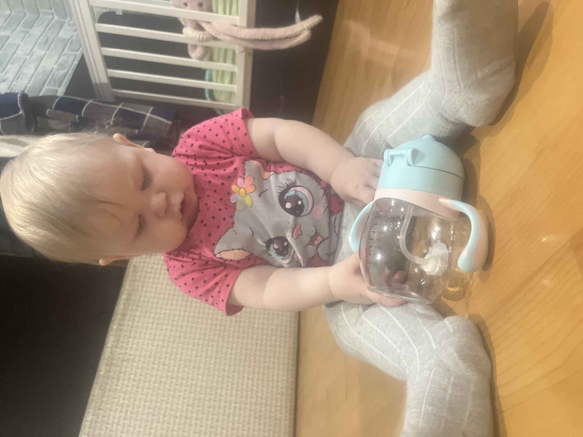 A baby with blonde hair is sitting on a wooden floor. The baby is wearing a pink shirt with a cartoon cat on it and grey and white striped leggings. The baby is holding a clear plastic bottle with a light blue top and handle. There is a white crib in the background with a blue blanket and a pink stuffed animal hanging from it. The location is unknown.