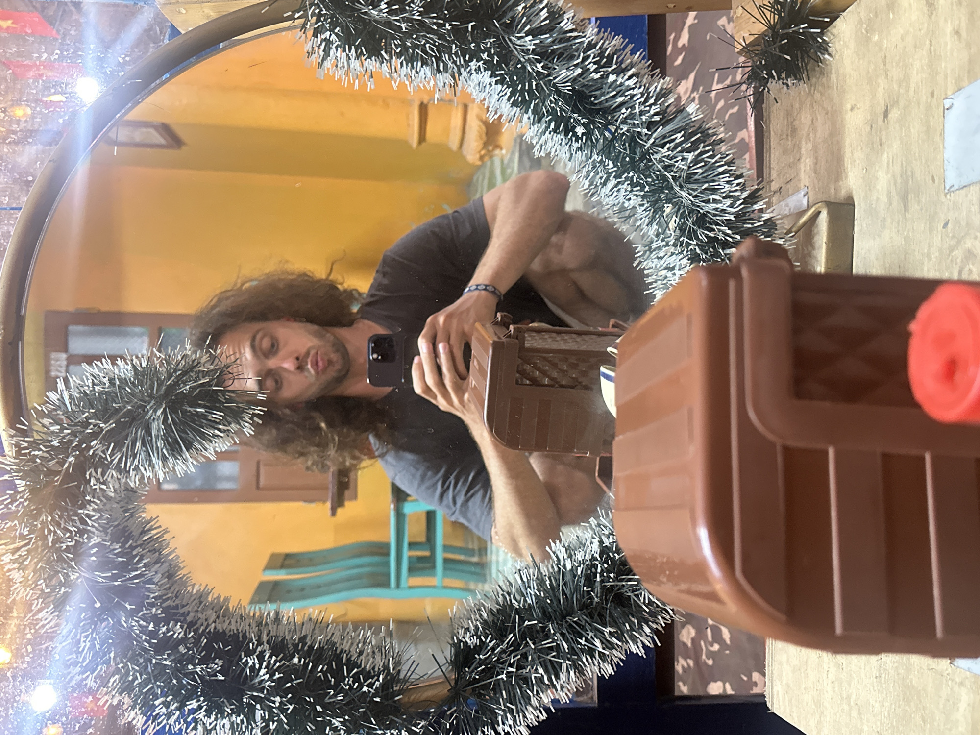 A person with long, curly, brown hair is captured in a selfie taken in a mirror. They are wearing a dark t-shirt and shorts, and are crouching down. The person is holding a smartphone in their right hand, appearing to take the picture. In the foreground, a brown, toy-like object resembling a vehicle or structure is visible. The mirror is adorned with a decorative, fluffy garland with white tips. The background, reflected in the mirror, shows a room with bright yellow walls, teal-colored chairs, and architectural elements like pillars and a wooden door. The scene appears to be indoors, possibly in a residential or commercial space. The location is identified as Tây Hoa Lư Ward, Vietnam.