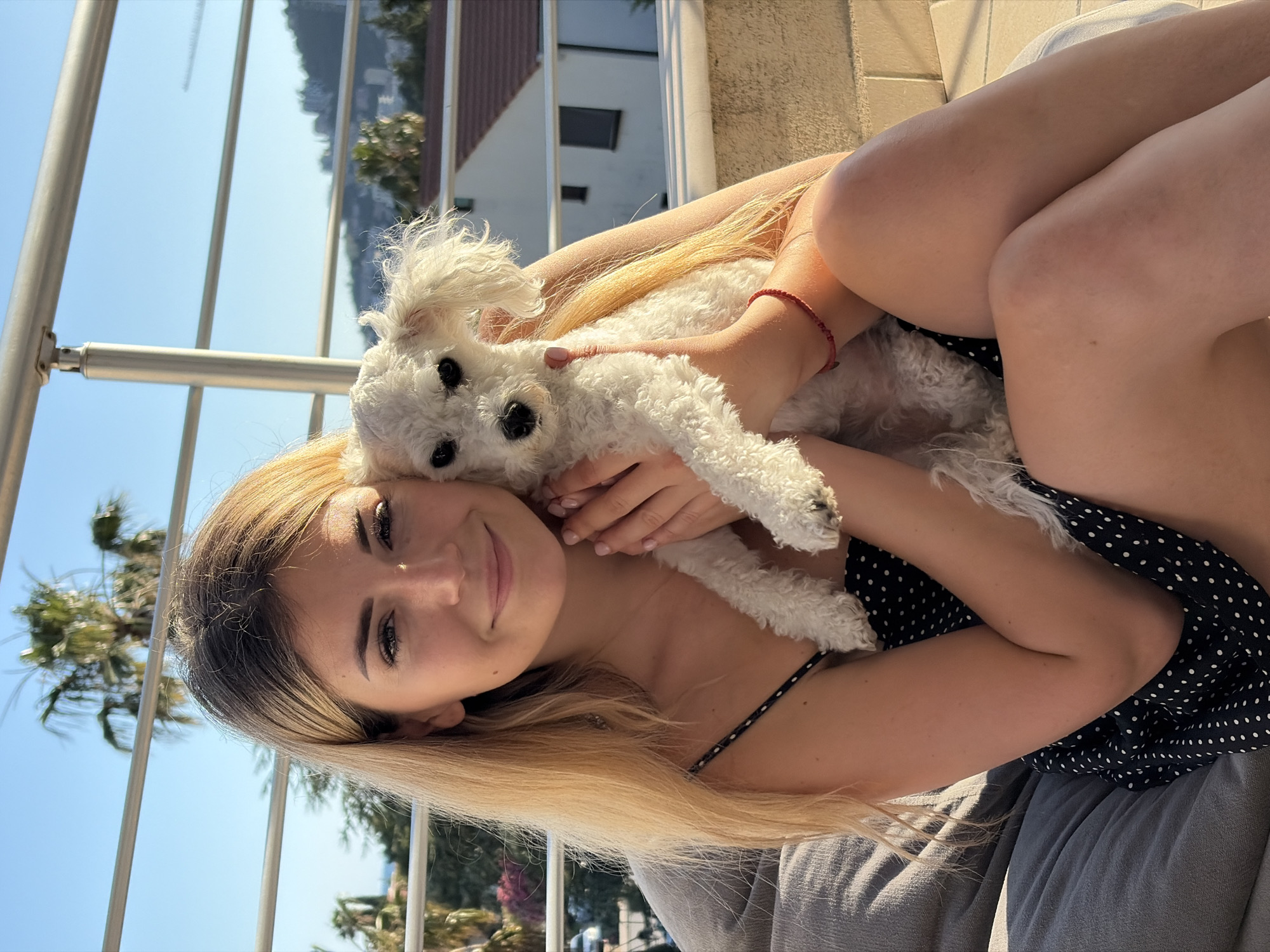 A young adult female with long blonde hair with darker roots smiles faintly while looking directly at the camera. She is seated on a grey cushion or beanbag chair, wearing a black polka-dot spaghetti strap dress. Her arms and bare legs are visible. A red string bracelet is on her right wrist. She cradles a small, white, curly-haired dog with dark eyes, which is looking slightly to the left of the camera.
The scene is set on a sunny outdoor balcony or terrace, indicated by a prominent silver metal railing with horizontal bars positioned behind the subjects. The background features a clear blue sky, several green palm trees, and typical Mediterranean-style white buildings with reddish-brown tiled roofs. In the distance, undulating green hills and mountains rise, and a glimpse of the blue Adriatic Sea is visible on the far left. The overall environment, characterized by the coastal setting, architecture, and mountainous backdrop, is consistent with the city of Budva, Montenegro. The warm, bright lighting suggests either morning or late afternoon sun.