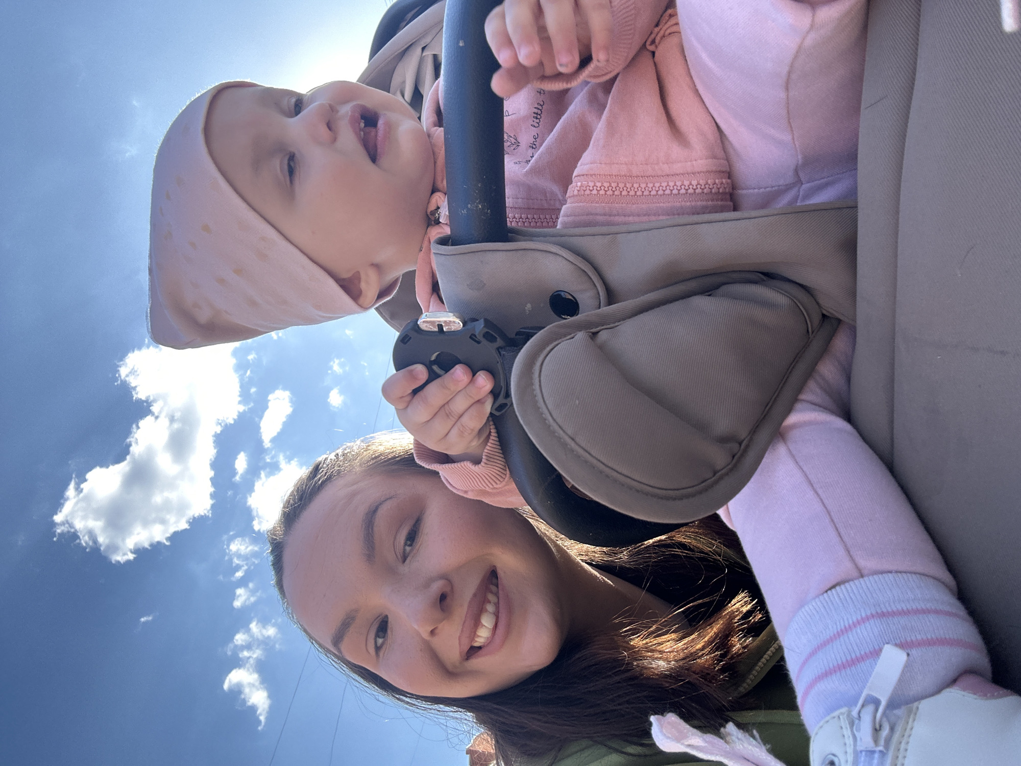 A low-angle, upward-facing shot captures a smiling woman and a baby in a stroller against a backdrop of a blue sky with scattered clouds. The woman has dark brown hair and is wearing an olive green jacket. The baby, positioned in the upper part of the frame, is wearing a pink outfit and a polka-dot bonnet. The stroller is gray and black. The background features a bright blue sky with white clouds. The image was taken outdoors on a sunny day. There is not enough information available to determine if this image was taken in the city of Chaikovsky, Russia.