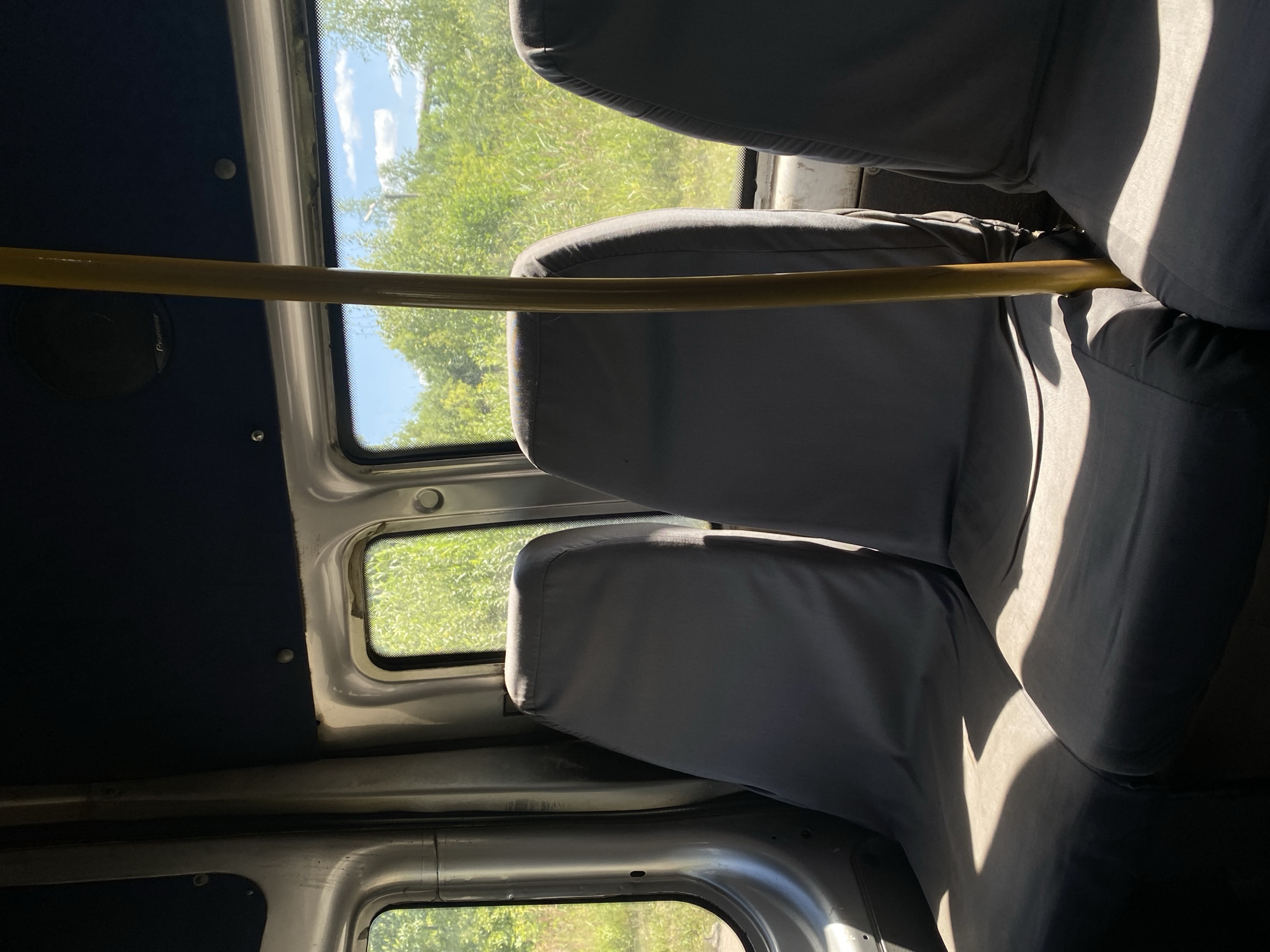 The image shows the interior of a passenger vehicle, likely a bus or van, with seats covered in dark grey fabric. Sunlight streams in through the windows, casting distinct shadows across the seats. A yellow pole, possibly a grab handle, stands vertically in the foreground between two seats. Outside the windows, lush green foliage and a blue sky with white clouds are visible, suggesting a rural or natural setting. The interior appears worn, with some of the metal framing and ceiling lining visible. The scene does not depict any people, specific city landmarks, or immediate actions. The location is stated to be Furmanov, Russia, though no visual cues within the image confirm this specific location.