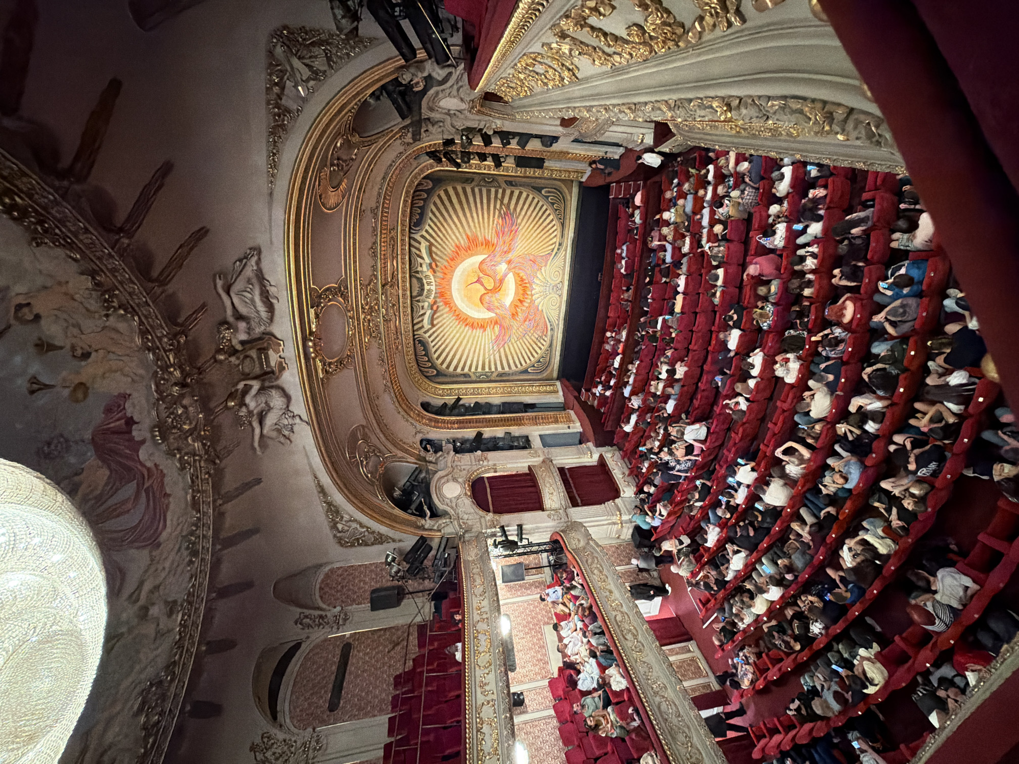 The image shows an interior view of a grand theater, likely in Sofia, Bulgaria, as indicated by the location context. The scene is filled with people seated in rows of plush red velvet chairs, facing a stage area that is not fully visible. The architecture of the theater is opulent, with gilded decorative elements and intricate artwork adorning the walls and ceiling.
The primary subjects are the audience members, who appear to be attending a performance or event. They are a diverse group, with many individuals looking towards the stage or engaged with their phones. The atmosphere seems to be one of anticipation or quiet engagement, typical of a theater setting.
Notable details include the ornate ceiling mural, which depicts a sun-like celestial body with birds or mythical creatures in flight, rendered in warm oranges, yellows, and reds. The elaborate gold detailing and moldings on the upper levels of the theater, as well as the draped red velvet curtains, contribute to the luxurious and historic ambiance. There is also a large, textured white light fixture visible on the left side of the frame, adding to the grandeur of the space. Lighting appears to be a mix of stage lighting and ambient illumination. The time of day is not explicitly discernible but the indoor setting suggests it could be evening or daytime during an indoor event.
There is some visible text on the ceiling mural, appearing as small, light-colored numerals and text within the sun-like graphic, though it is too small and blurry to be definitively read. The overall impression is that of a lively cultural event taking place within a beautifully preserved historic building.