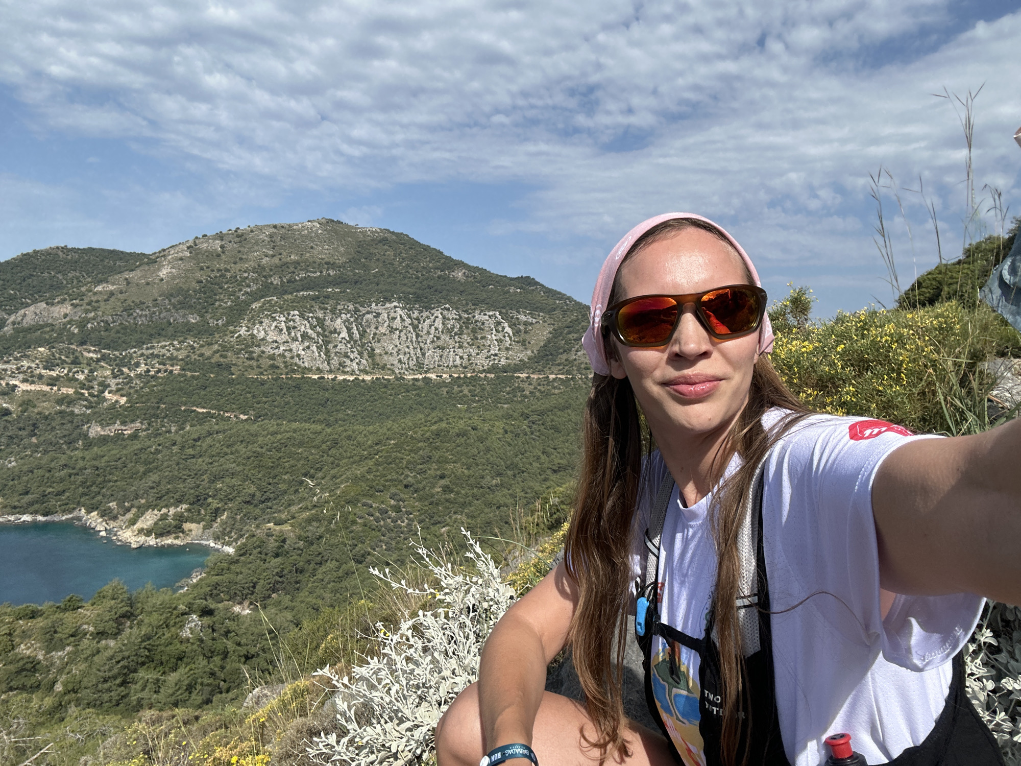 A person with long brown hair, wearing sunglasses and a pink headscarf, is seated and taking a selfie. The background features a mountainous landscape with dense green vegetation and a body of water. The location is likely Fethiye, Turkey, given the typical Mediterranean scenery with rugged hills and a turquoise coastline. The sky is partly cloudy. The person's attire suggests outdoor activity, possibly hiking or running. The vegetation in the foreground is sparse, consisting of low-lying plants and shrubs.