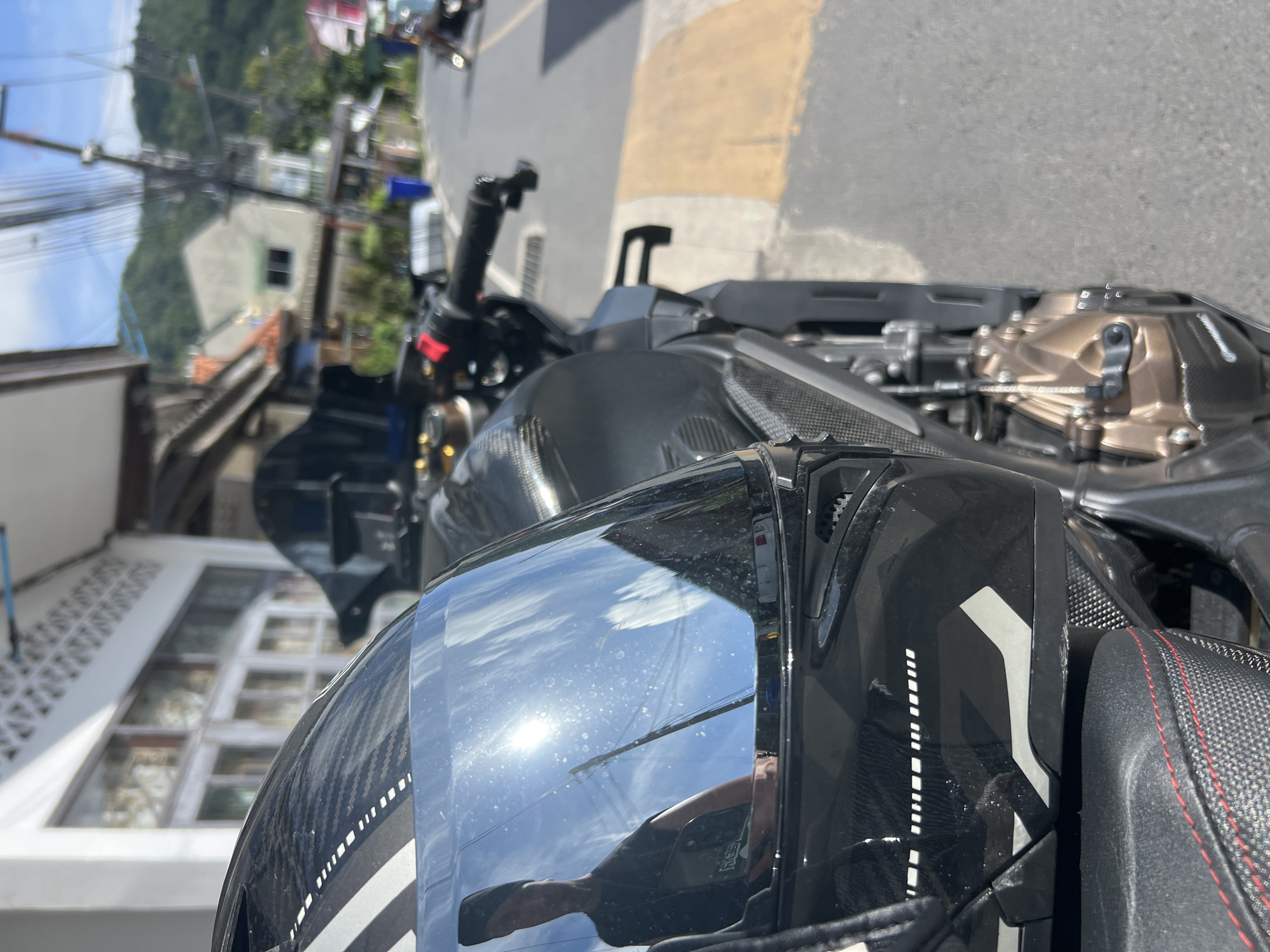 In Choeng Thale, Thailand, a black motorcycle is parked on the side of a street. A black helmet with a reflective visor is placed on the seat of the motorcycle, its visor reflecting the sunny sky, clouds, and a portion of a person wearing a black t-shirt. The motorcycle features a carbon fiber design on its side panel and a bronze-colored engine casing. In the background, buildings with windows and a blue trash bin are visible along the street, with power lines and lush green hills in the distance.