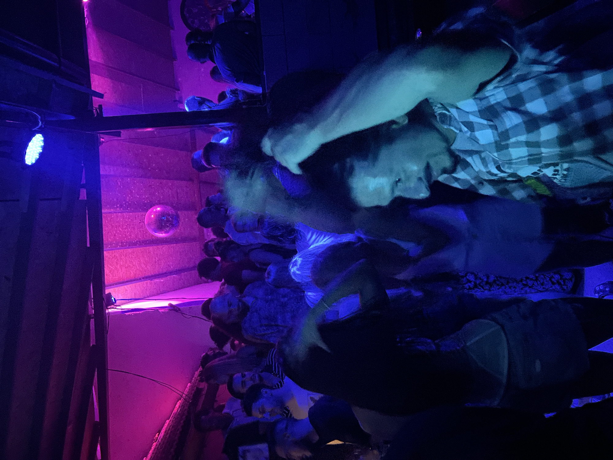 The image depicts a crowded indoor nightclub scene illuminated by vibrant pink and blue lights. A disco ball is visible hanging from the ceiling. In the foreground, a man with long dark hair and a mustache, wearing a plaid shirt, is looking to his left while raising his arms. Several other individuals are present, some dancing and interacting, while others are looking in various directions. The environment appears to be a dimly lit, energetic venue, likely a bar or club, located in Herceg Novi, Montenegro.