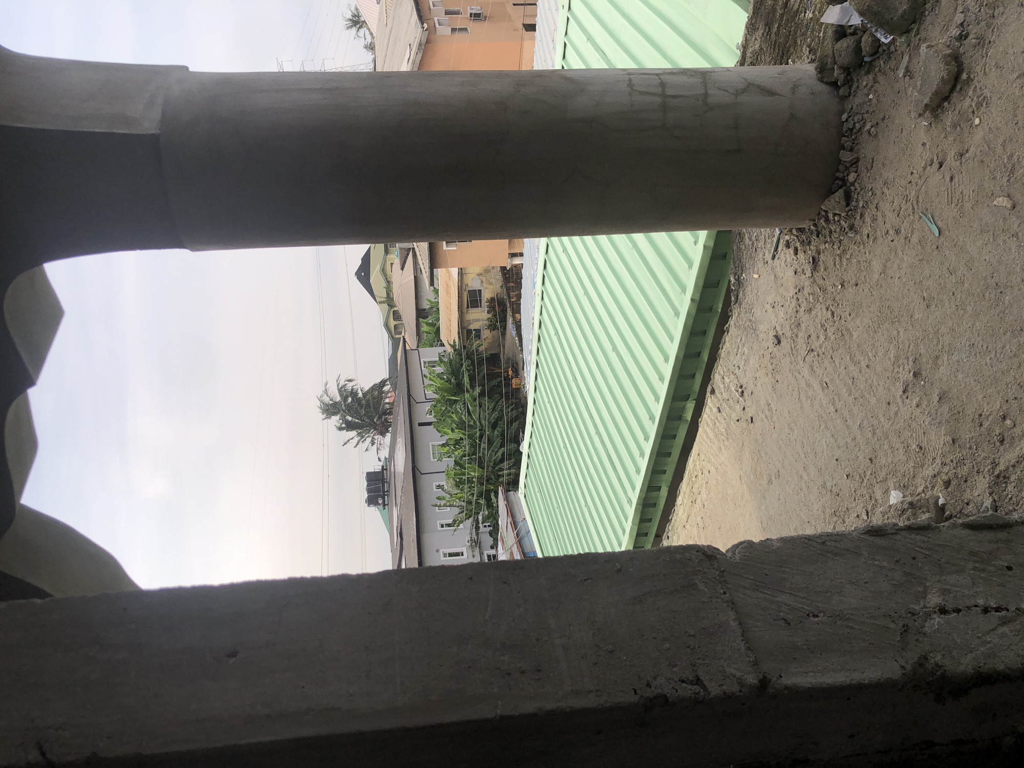 This image depicts an exterior view of buildings and construction work in Ado, Nigeria. In the foreground, a large concrete pillar and a section of a rough concrete wall frame the scene. Beyond this, a vibrant green corrugated metal roof covers a structure, possibly a container or a low-rise building. Further in the background, several multi-story residential buildings are visible, characterized by their varied architectural styles and pastel colors, including an orange and a pale gray facade. Some buildings feature air conditioning units and water tanks on their roofs. Lush green vegetation, including what appears to be banana trees and palm trees, is interspersed among the buildings. The sky is overcast, suggesting a cloudy day. The ground in the foreground is covered with loose soil and small debris. There are no people visible in the image.