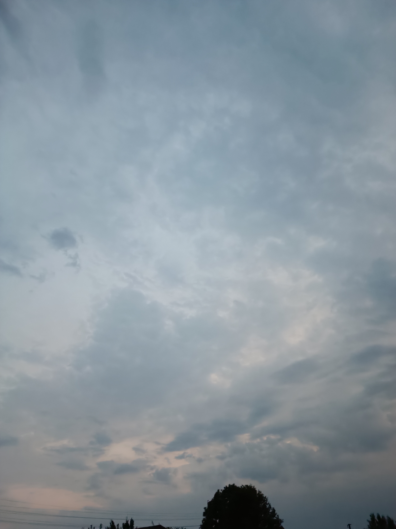 This image captures a dusky sky filled with scattered clouds, casting a muted light. The lower portion of the frame reveals a silhouetted treeline and what appears to be the roof of a building. Utility wires are also visible stretching across the bottom of the image, suggesting an urban or suburban environment. No people are present, and no specific actions or interactions are depicted. The overall atmosphere is subdued, likely captured during twilight or a heavily overcast day.
