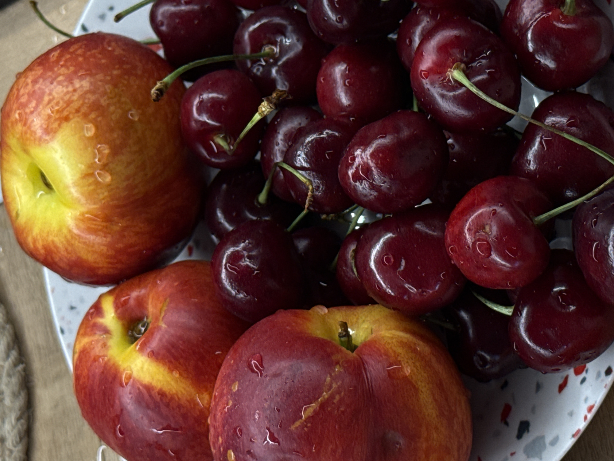 A close-up, high-angle shot captures a vibrant still life arrangement of fresh fruits on a white speckled plate. The primary subjects are three reddish-yellow peaches or nectarines and a generous pile of dark red cherries, all glistening with water droplets.
The peaches/nectarines, positioned at the bottom left and right of the frame, exhibit a smooth skin texture with prominent red and yellow hues, showing faint streaks and a natural indentation at their tops. Numerous small water droplets cling to their surfaces, reflecting light.
Interspersed and piled above the peaches/nectarines are many dark, glossy cherries, still attached to their green stems. Their deep maroon color contrasts with the brighter tones of the other fruits. Like the peaches, these cherries are adorned with clear water droplets, indicating freshness or recent washing.
The fruits rest on a white ceramic plate, which has a distinctive speckled pattern of small red, grey, and black flecks. Part of a rustic, woven or braided item (possibly a placemat or basket made of natural fibers) is visible in the blurred background to the left, resting on a light brown wooden surface. There is no visible text in the image. The scene conveys a sense of natural abundance and freshness, likely taken indoors in Pervomaysky, Russia.