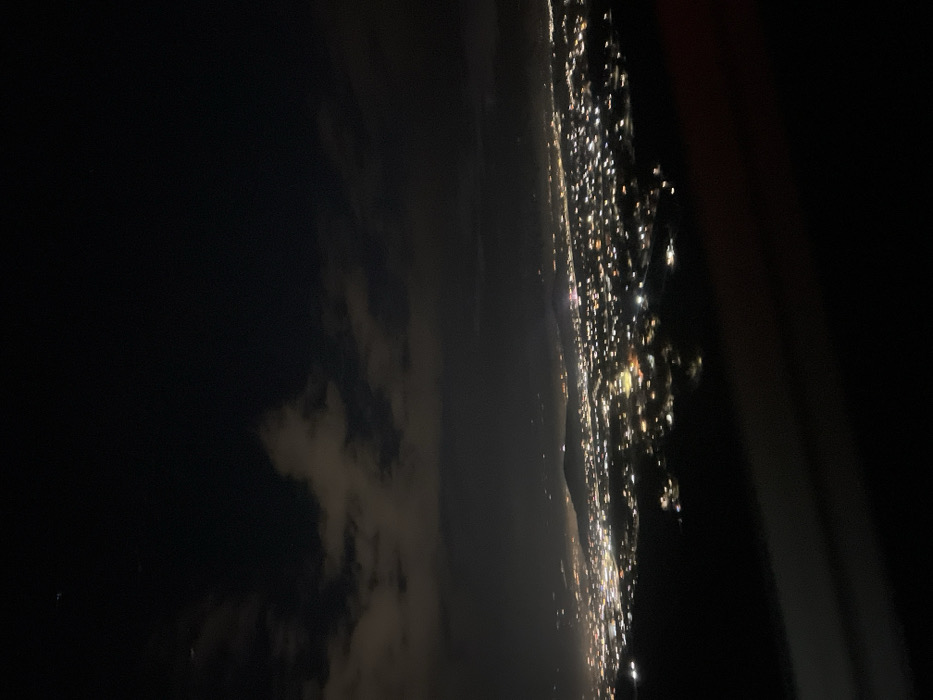 An aerial nightscape reveals a coastline speckled with bright city lights. The dense urban glow contrasts with the dark expanse of the ocean or sky. Some cloud cover is visible on the left. Part of the plane's interior frame can be seen on the right of the image.