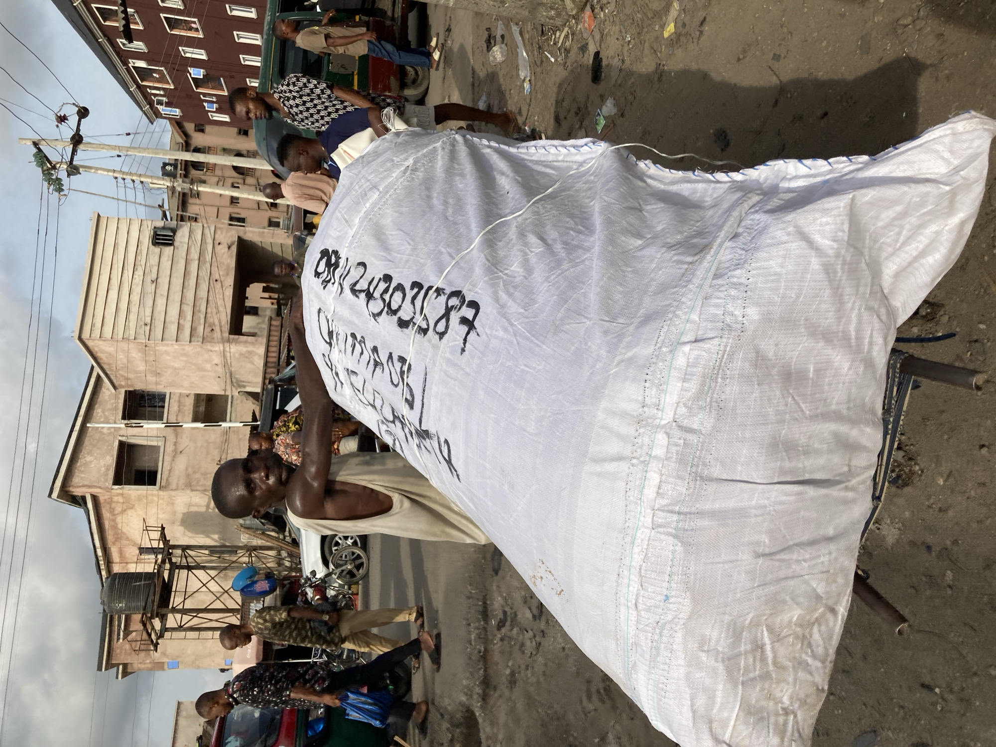 A street scene in Ura Nta Umuarandu, Nigeria, is captured. A man with dark skin and a sleeveless top strains to lift a large white sack onto a bicycle. The sack has a phone number written on it. Several other people are present in the background, standing near vehicles and buildings. The environment appears to be urban, with buildings, utility poles, and evidence of foot traffic.