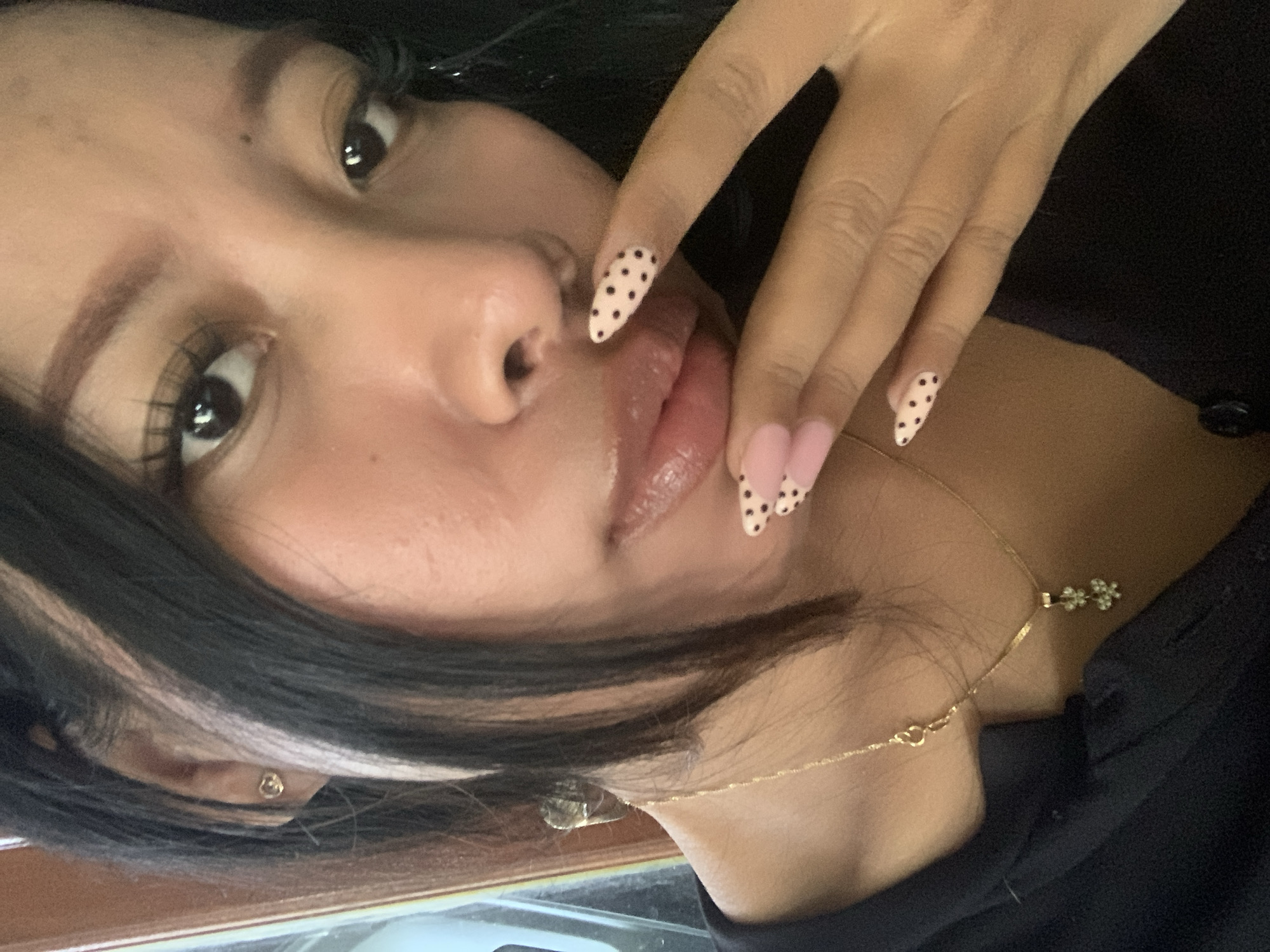 A close-up, chest-up, indoor portrait shows a young woman with dark hair and fair skin. She is wearing a black shirt and a gold necklace with a pendant shaped like two flowers. Her nails are decorated with a pink French manicure with black polka dots. She is touching her lips with her right hand. The background is out of focus, but appears to be a room with a wooden door frame. There is no visible indication of the city of Jajag, Indonesia.