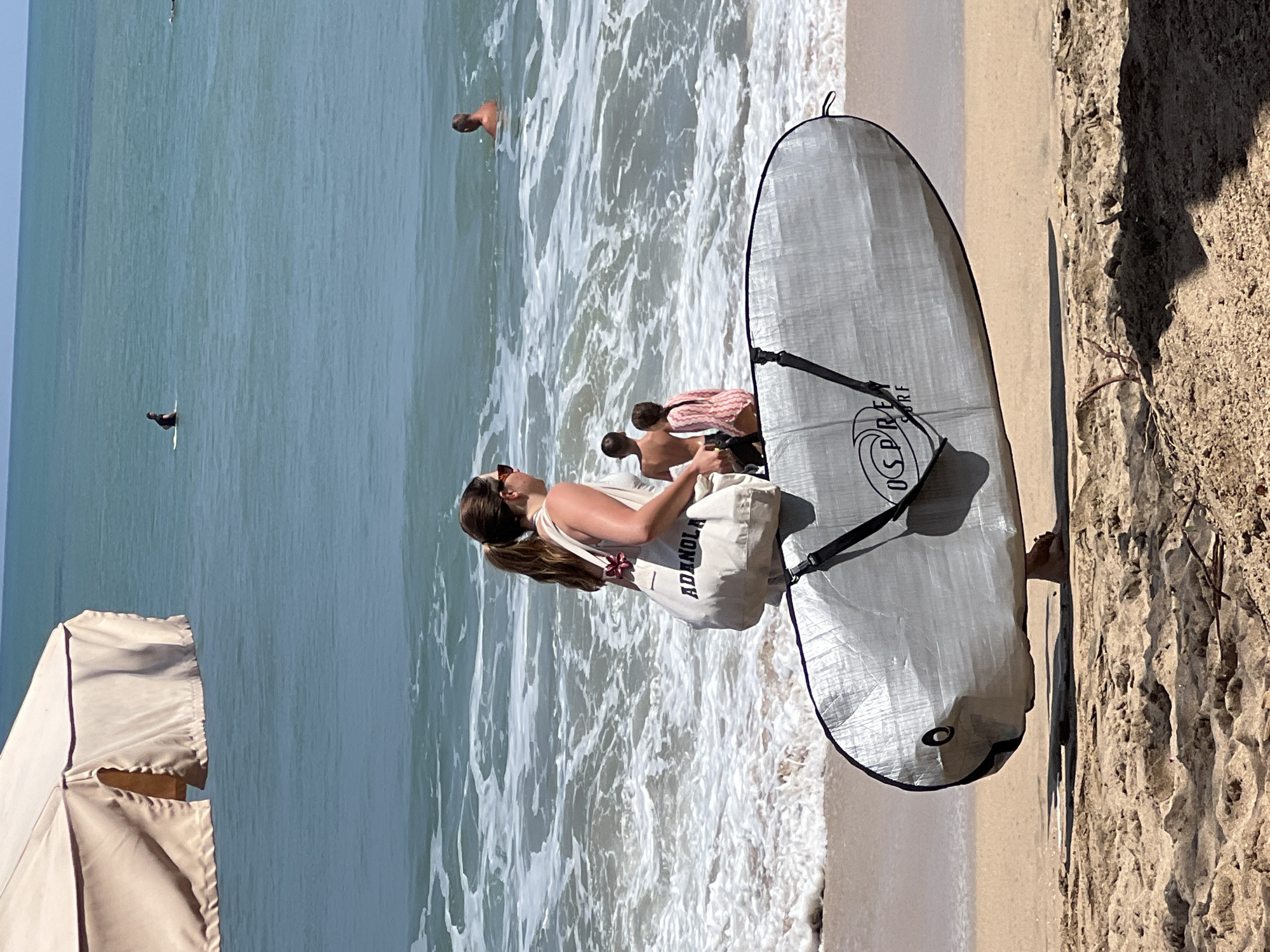 A woman with long brown hair pulled into a ponytail, wearing sunglasses and a white top, stands on a sandy beach in Kuta, Indonesia. She is carrying a large tan tote bag with the word "ADANOLA" printed on it, and a gray surfboard bag is held in front of her. The surfboard bag has the Osprey Surf logo. In the background, the turquoise ocean has white waves crashing near the shore. Several people are visible in the water; one man is sitting on a surfboard, another man is partially submerged, and two children, a boy and a girl in a pink and white striped outfit, stand in the shallow water. A beige beach umbrella is partially visible in the upper left corner of the frame. The sand in the foreground is textured with shadows and a few small pieces of dried vegetation.