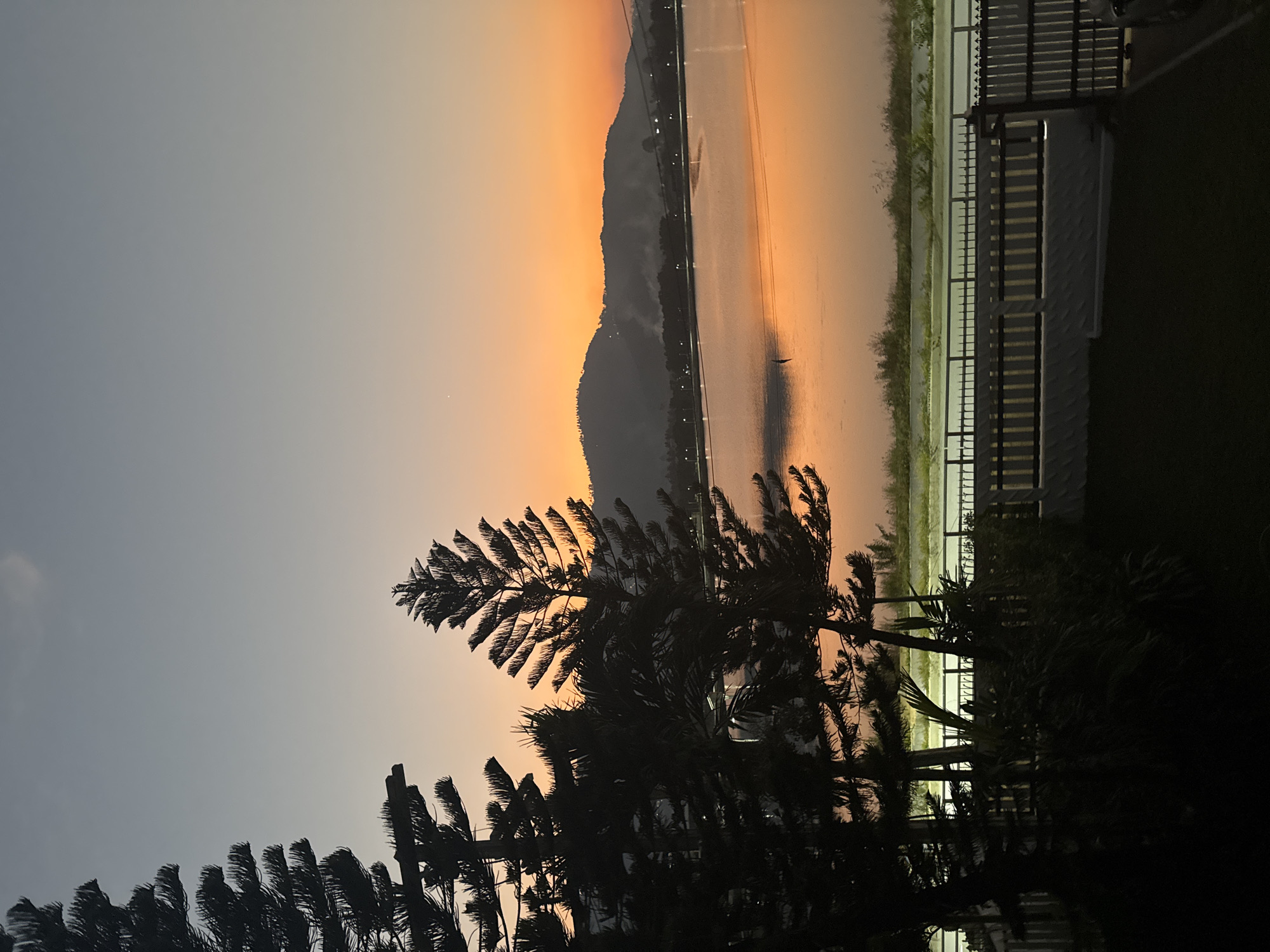 The image depicts a sunset over a body of water with silhouetted trees in the foreground. The sky is a gradient of orange and grey. In the middle ground, a mountain range is visible along with the opposite shore of the body of water. A fence is located along the shore. The scene appears to be in or near Baan Chaweng, Thailand based on the geographical characteristics. A solitary figure is on the water.