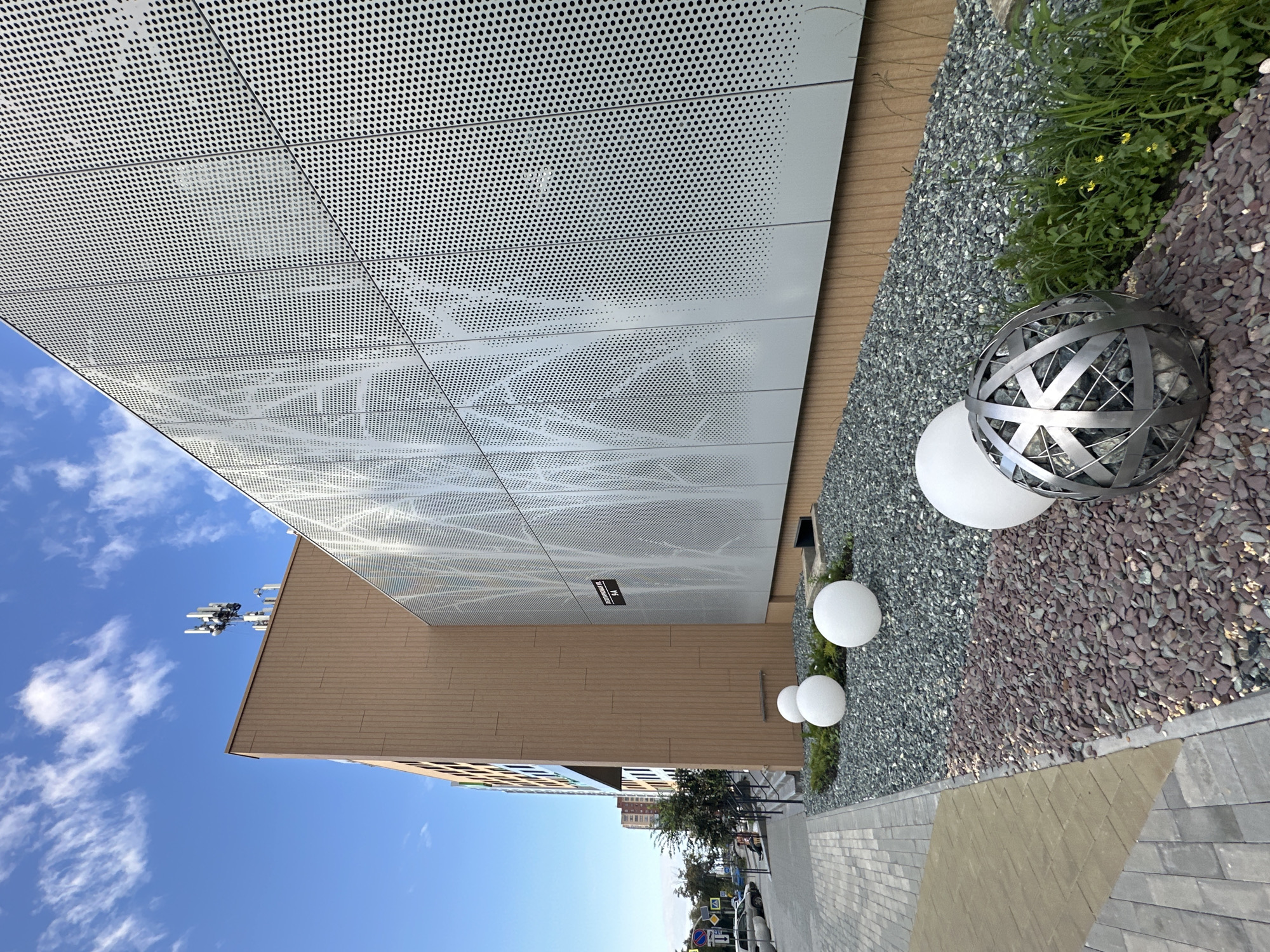 An exterior, low-angle shot captures a modern building with a brown facade and a silver perforated metal exterior panel. The silver panel features a pattern of small circles and a subtle tree-like design. On top of the brown section of the building, a telecommunications antenna is visible against a bright blue sky with wispy white clouds. In the foreground, a paved walkway with geometric patterns transitions to a landscaped area. This area is covered in grey and reddish-brown gravel and features several large white spherical lights and a decorative metal sphere filled with stones. A street with parked cars and trees is visible to the left of the building, indicating an urban environment. A street sign with the number "9A" is mounted on the silver panel of the building.