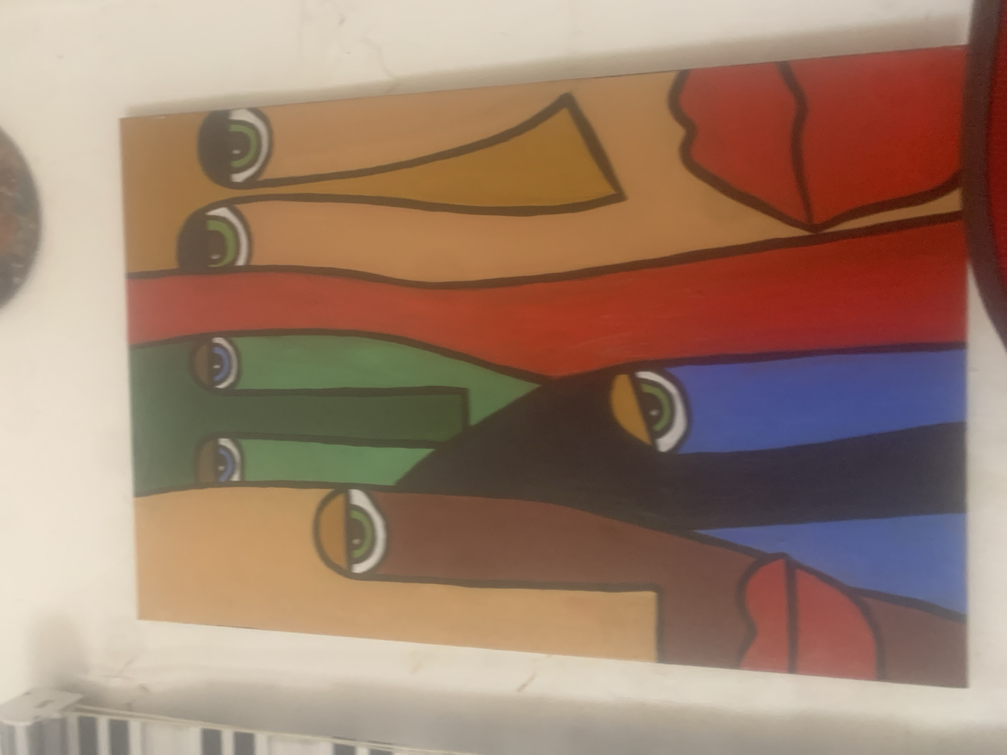 The image displays a square abstract painting featuring a mosaic of stylized faces. The faces are rendered with bold, flat colors and sharp geometric shapes, with overlapping elements creating a sense of depth and complexity. Prominent features include large, almond-shaped eyes with distinct irises and pupils, as well as full, red lips. The color palette consists of earthy tones like beige, brown, and orange, contrasted with vibrant green, red, and blue. The painting is hung on a plain white wall, with the top edge of the artwork visible. To the left of the painting, a portion of what appears to be a window blind with horizontal slats is visible. The lighting suggests an indoor setting. The provided context indicates this artwork is located in Ado, Nigeria.