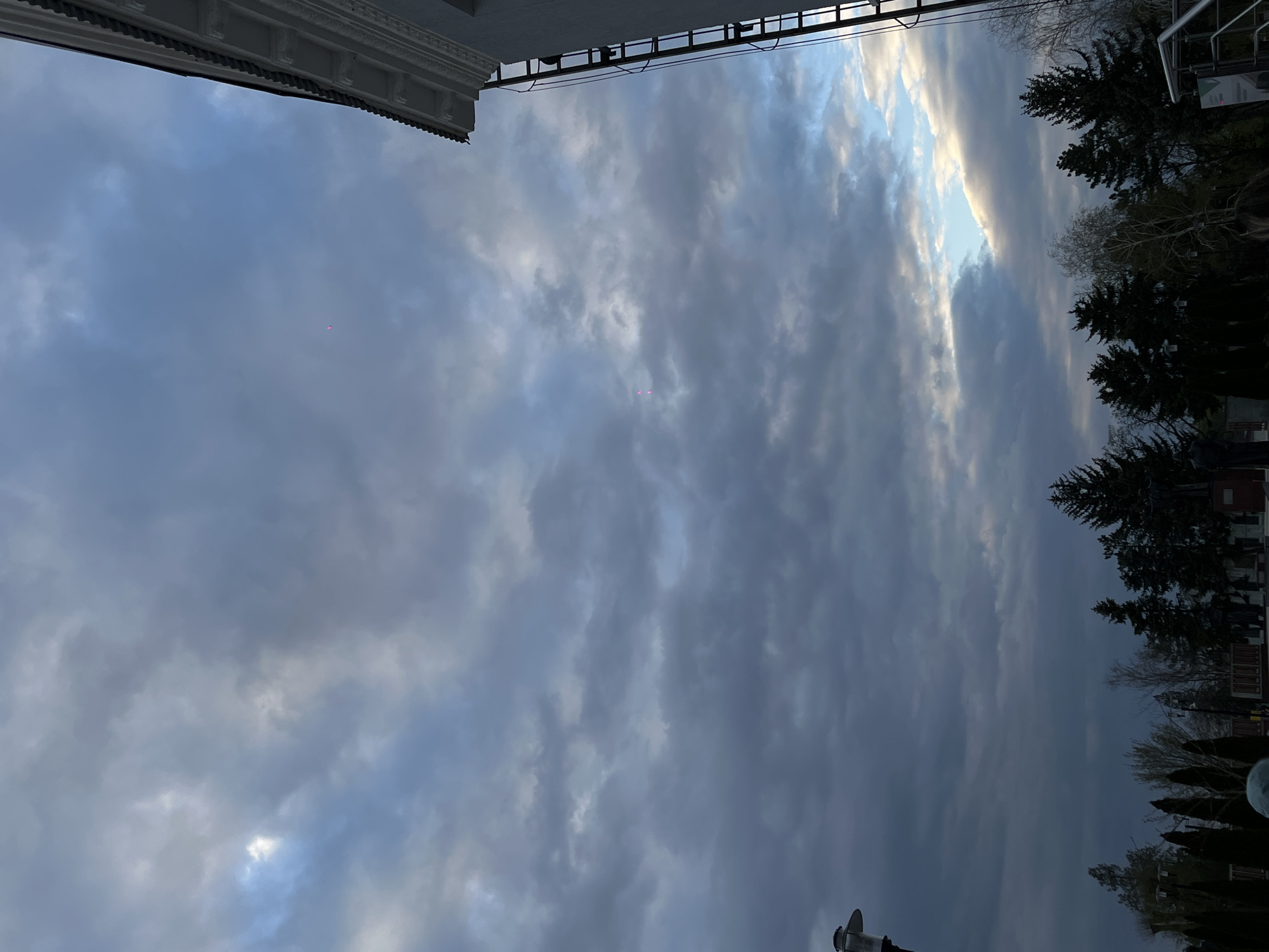 The image, taken from a low angle, features a cloudy sky in Novopokrovskaya, Russia. The upper portion of a building's architectural details is visible at the top left, while trees and some structures suggest a street scene on the right. The sky dominates the view, with varying cloud formations and a small opening hinting at sunlight. Two pink dots are noticeable in the sky, possibly from a light source.