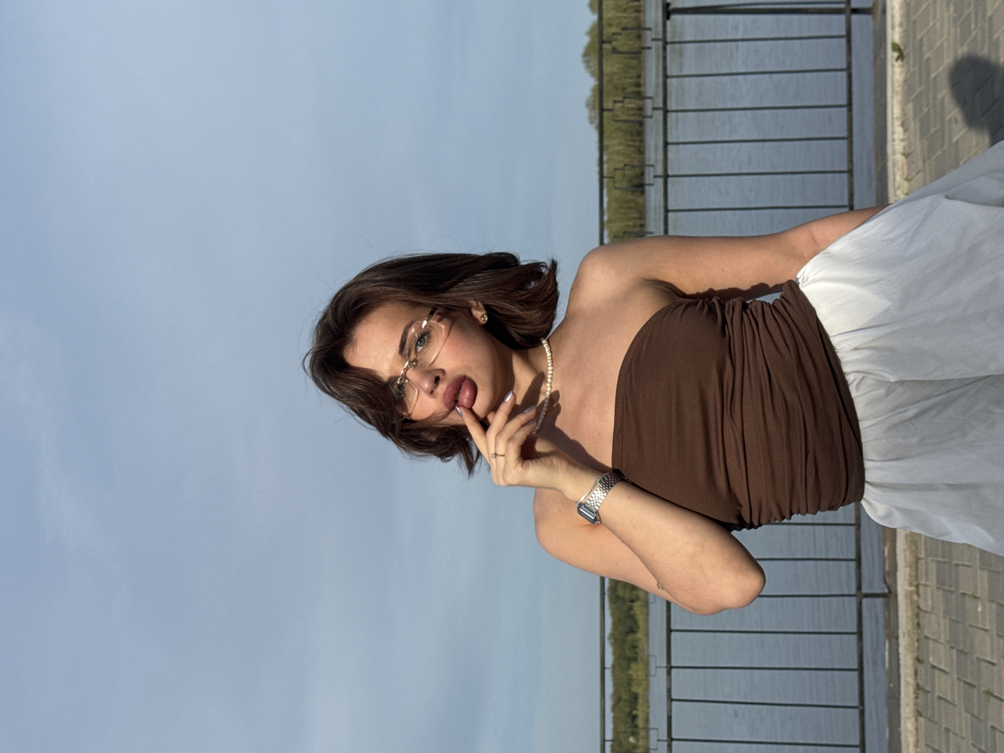 A woman stands in front of a railing in Sarapul, Russia, likely near a body of water. She wears a brown strapless top, a white skirt, glasses, and a watch. Her hair is short and brown. She is holding a finger to her lips. The background includes a blue sky, water, and green vegetation on the opposite shore.