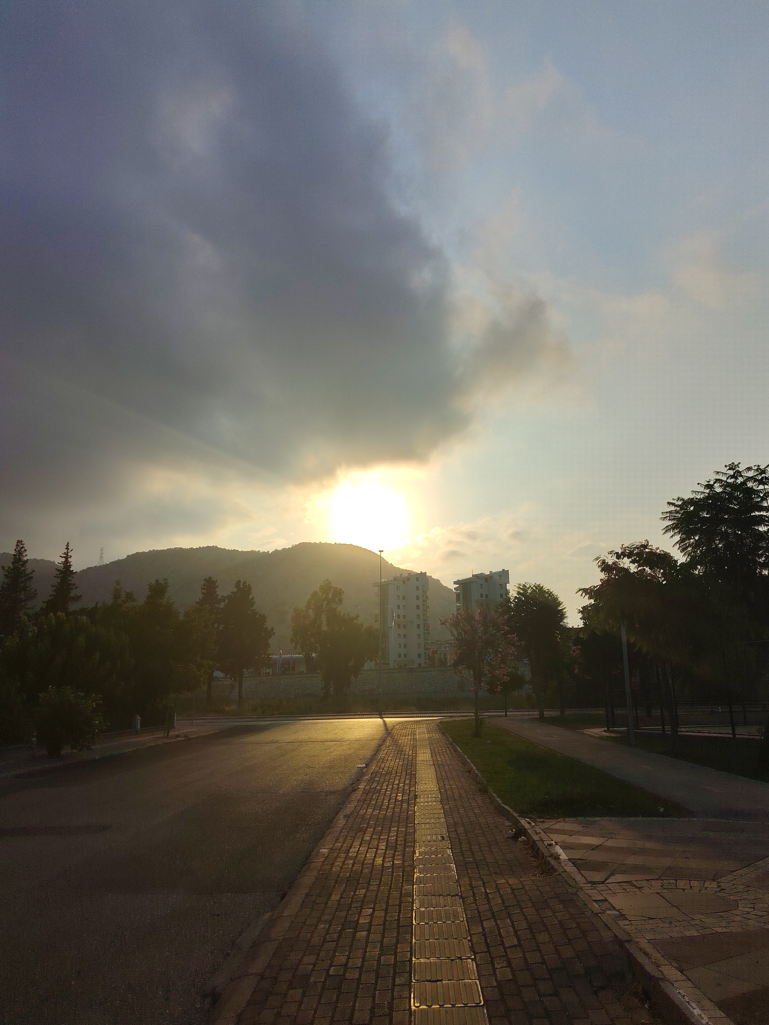 The photograph captures a street scene in Konyaaltı, Turkey, during what appears to be sunrise or sunset. The sky is dramatic, with heavy, dark clouds on the left partially obscuring a bright sun peeking through on the right, casting rays of light. In the background, a low, tree-covered hill is visible, with a few tall buildings situated on its slopes. In the middle ground, there are more trees and some modern, multi-story buildings. In the foreground, a paved road runs diagonally from the lower left towards the upper right. To the right of the road, a brick walkway with a tactile paving strip is prominent, leading towards the viewer. The light from the sun reflects strongly on the wet surface of the road and the brick walkway, creating a golden glow. The overall impression is a serene, possibly urban landscape at the beginning or end of the day. There are no people or animals visible in the image.