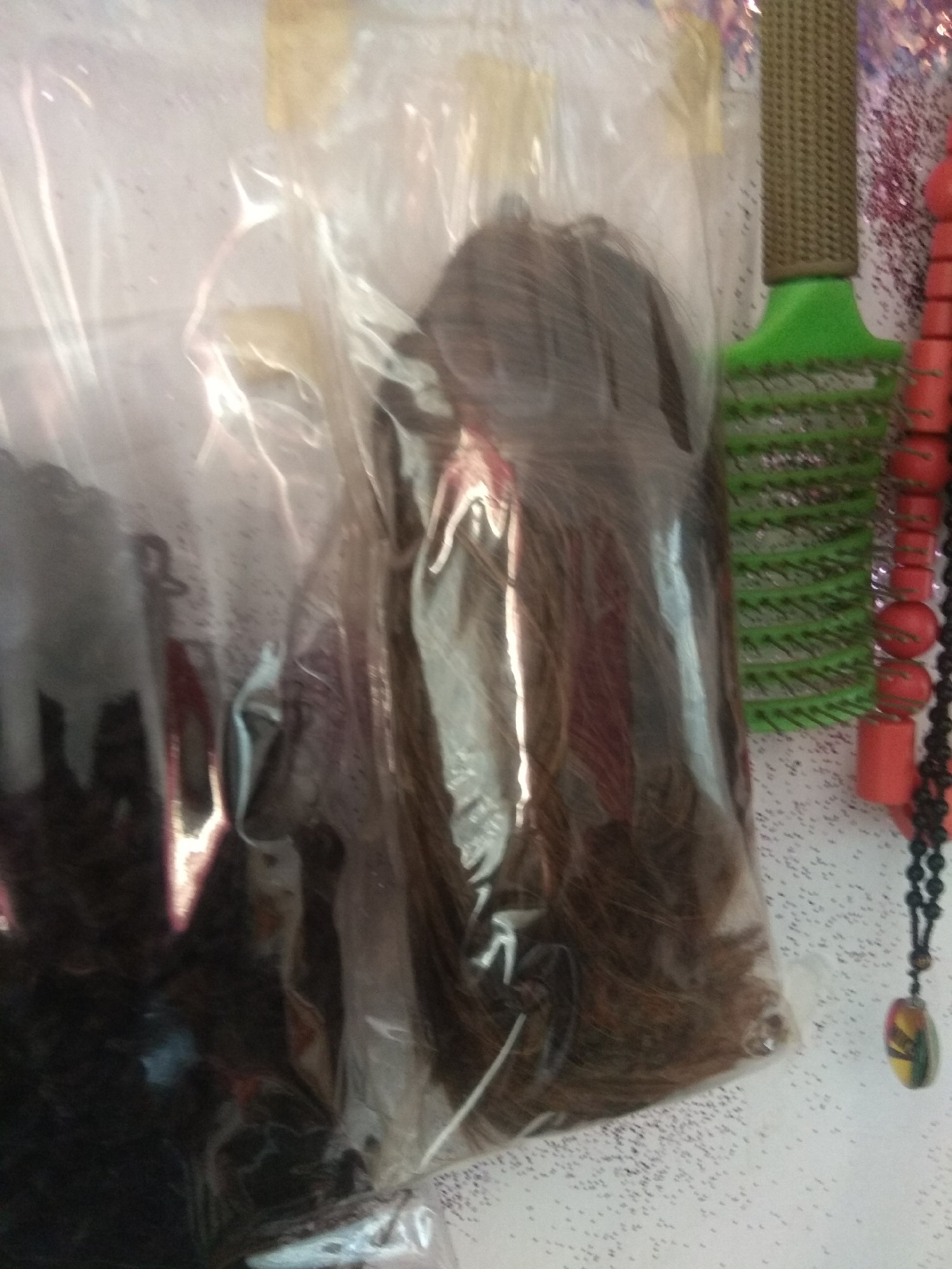 The image displays a close-up view of a plastic bag containing dark, brown hair, possibly extensions or a wig. The bag is partially transparent, allowing the texture and color of the hair to be seen. To the right of the bag, a green hairbrush with a brown handle is visible. Further to the right, a string of red, cylindrical beads and a black beaded necklace with a pendant featuring an abstract design are present. The background appears to be a textured surface with scattered small dark specks. The lighting creates highlights and shadows on the plastic bag and its contents. The location is stated to be in the city of Jalingo, Nigeria.