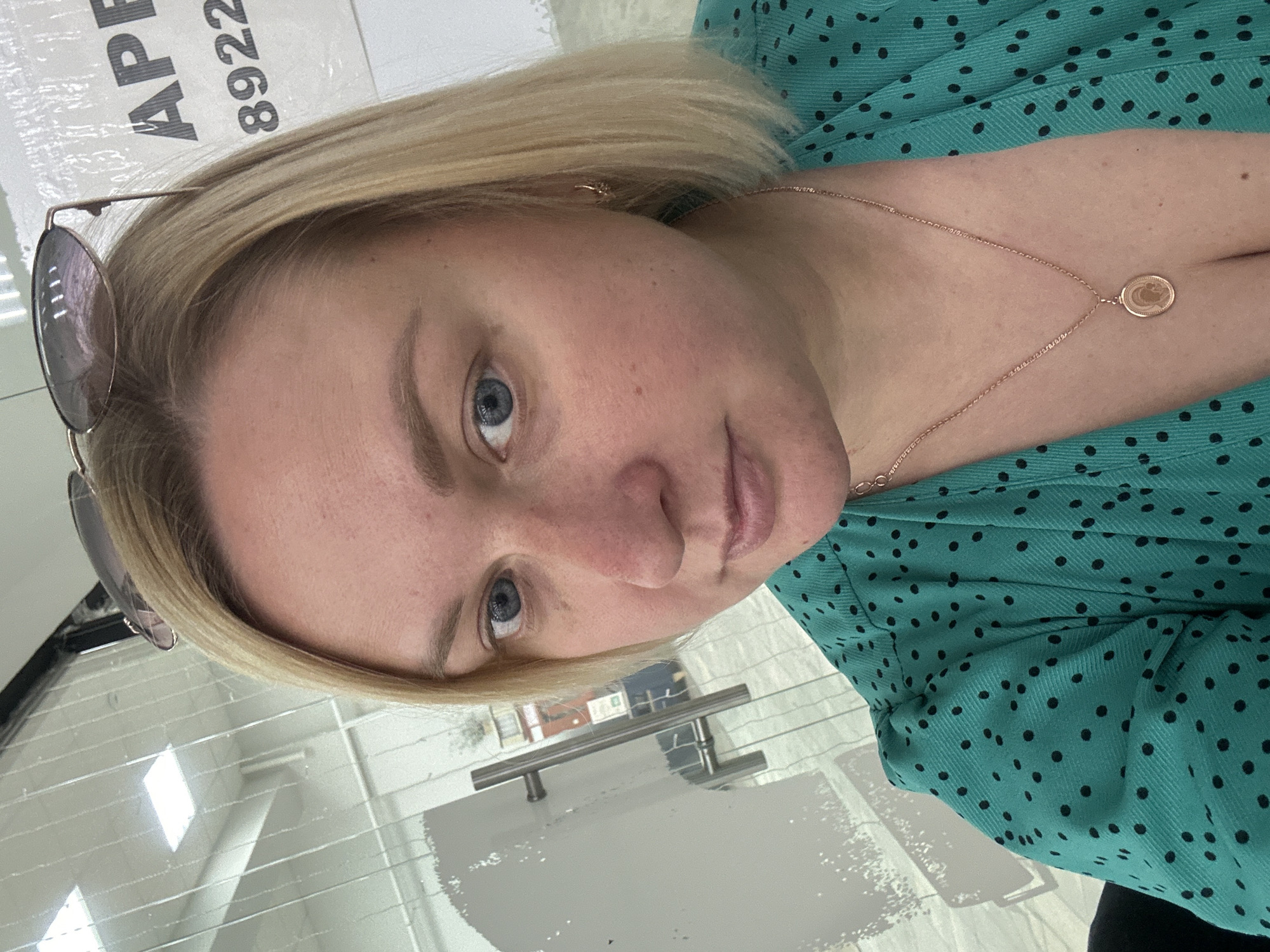In the city of Chaikovsky, Russia, a woman with blonde hair and blue eyes, wearing a green polka-dot dress and a rose gold necklace, is taking a selfie. Her sunglasses are perched on top of her head. The background appears to be an office environment with glass walls and doors, and a sign with the word "APE" and the number "8922" is visible.