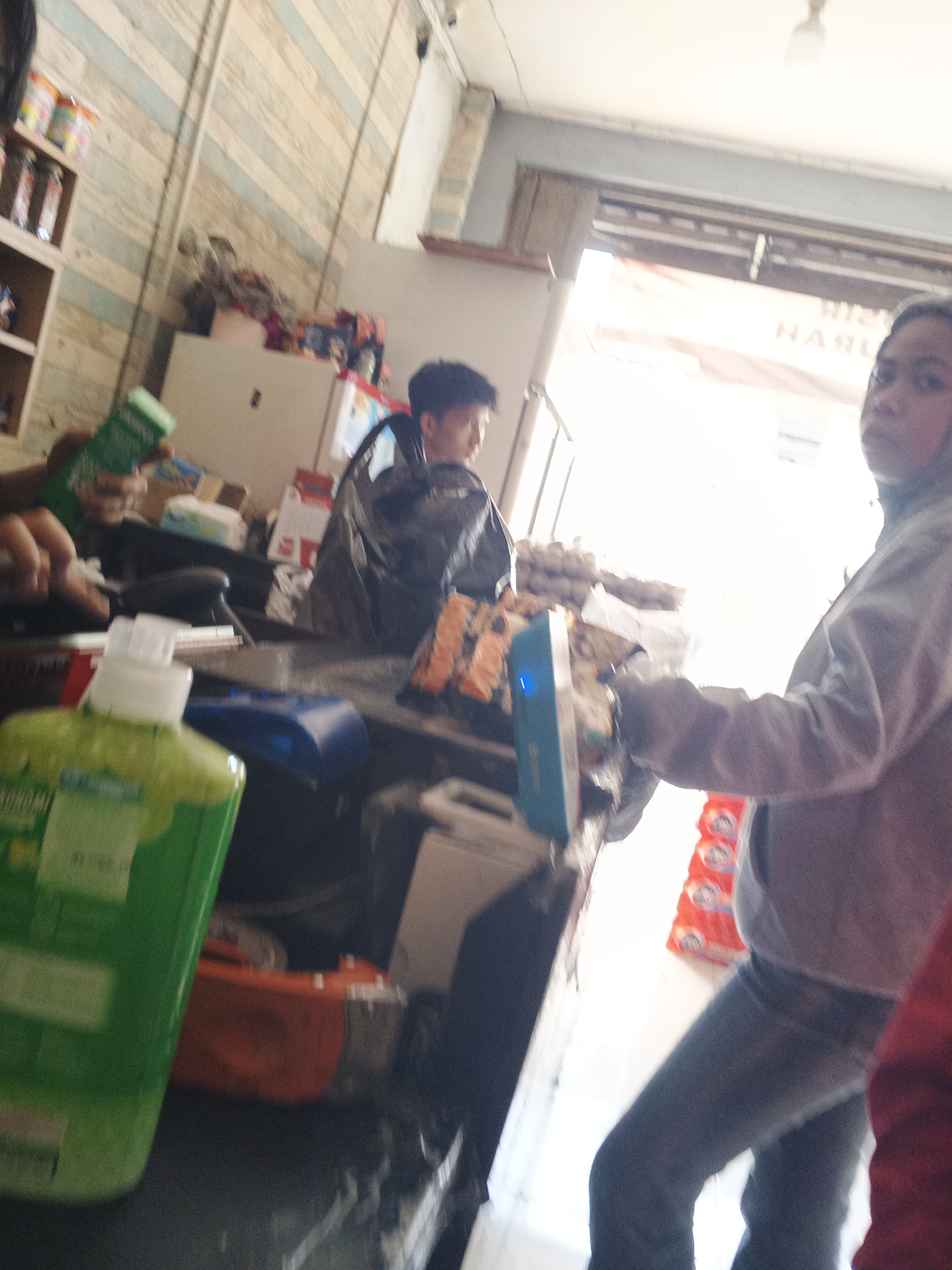 The image depicts an indoor scene, likely a retail establishment, with an unfocused view of people and merchandise. In the foreground, a green bottle of liquid, possibly soap or detergent, is prominently displayed on a counter. To the left, a person's hand is visible, holding a green bottle. In the middle ground, another person is seen wearing a dark jacket, and further back, a white appliance, perhaps a refrigerator or cabinet, is present. The background reveals a doorway leading to a bright area, with text on a sign that is partially obscured and difficult to read, but appears to contain the word "NARU". In the right portion of the image, a person in a gray hoodie and jeans is visible, seemingly interacting with a blue electronic device on the counter. Stacked red packaging with white circles and figures is also visible to the right. The overall environment appears cluttered and somewhat disorganized. The location in the city of Sukabumi, Indonesia is not discernible from the visual information provided in the image.