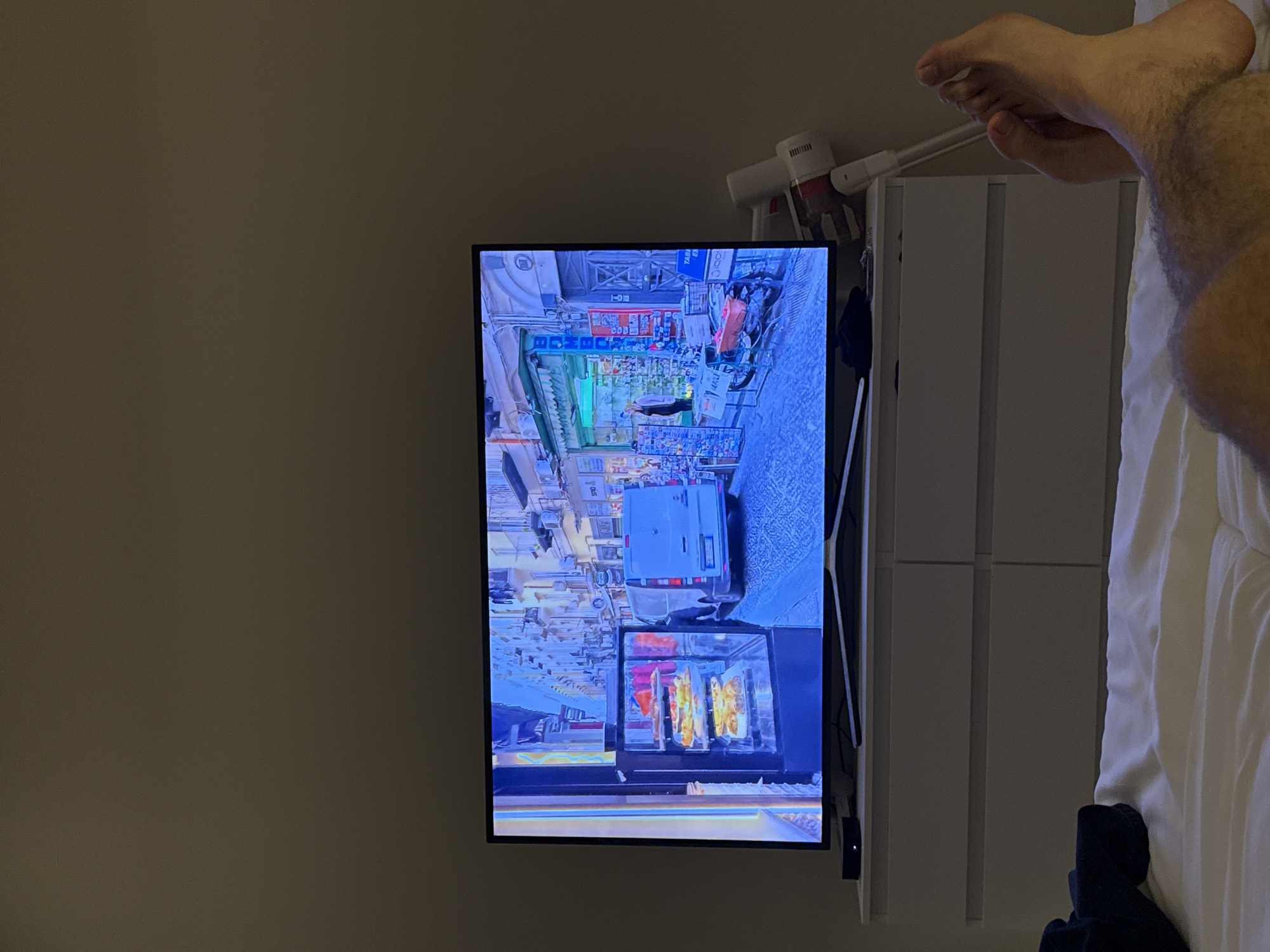 This media file shows a person's foot and leg resting on a white comforter. The person is likely in a bedroom, given the presence of a bed and what appears to be a dresser. A television is mounted on the wall to the left of the dresser, displaying a colorful image of a busy street market scene. The scene on the TV appears to be a bustling outdoor market with stalls and people. Visible text on the TV screen includes signs with what looks like Asian characters. To the right of the TV, a white cordless vacuum cleaner is leaning against the dresser. The lighting suggests it could be evening or a dimly lit room. The overall setting is domestic and relaxed, with the person seemingly unwinding and watching TV. The location context of Dubai, United Arab Emirates, doesn't offer specific visual cues within the frame.