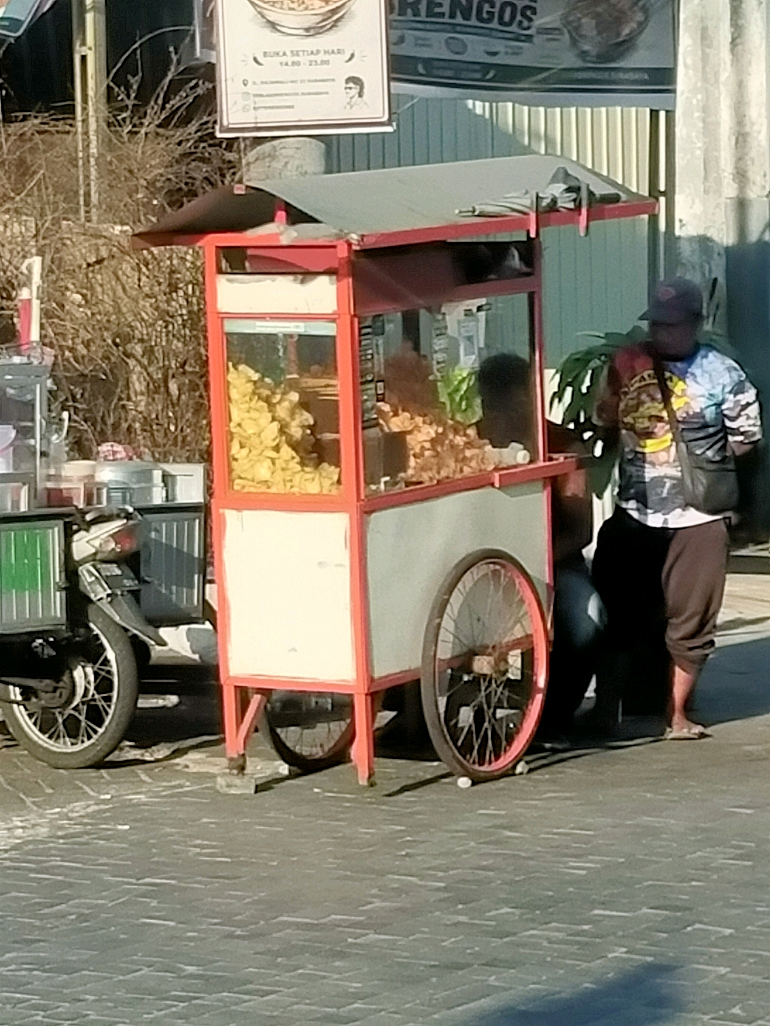 In Surabaya, Indonesia, a street vendor is seen standing beside a mobile food cart. The cart is painted red and white, with a glass display case filled with food. A motorcycle is parked to the left of the cart. The vendor is wearing a colorful t-shirt and dark pants. The scene appears to be taking place on a paved street, with a building in the background. The lighting suggests it is daytime.