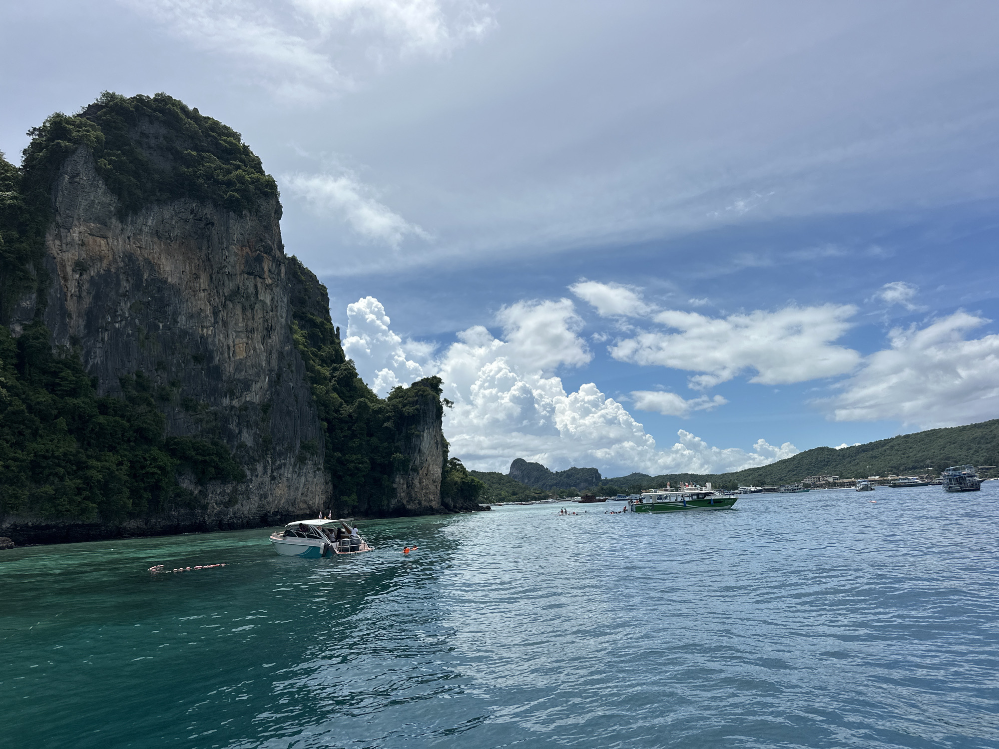 The image depicts a scenic coastal view in Ao Nang, Thailand, characterized by a towering limestone cliff on the left, lush green vegetation covering its slopes, and a turquoise sea. Several boats are present in the water. In the foreground, a white and blue boat with passengers is visible. Further out in the water, a line of people appear to be swimming or floating, possibly part of a tour group. Another green and white boat is seen in the middle ground, also with passengers. In the background, more boats and the coastline of Ao Nang, dotted with buildings and greenery, are visible under a partly cloudy blue sky.