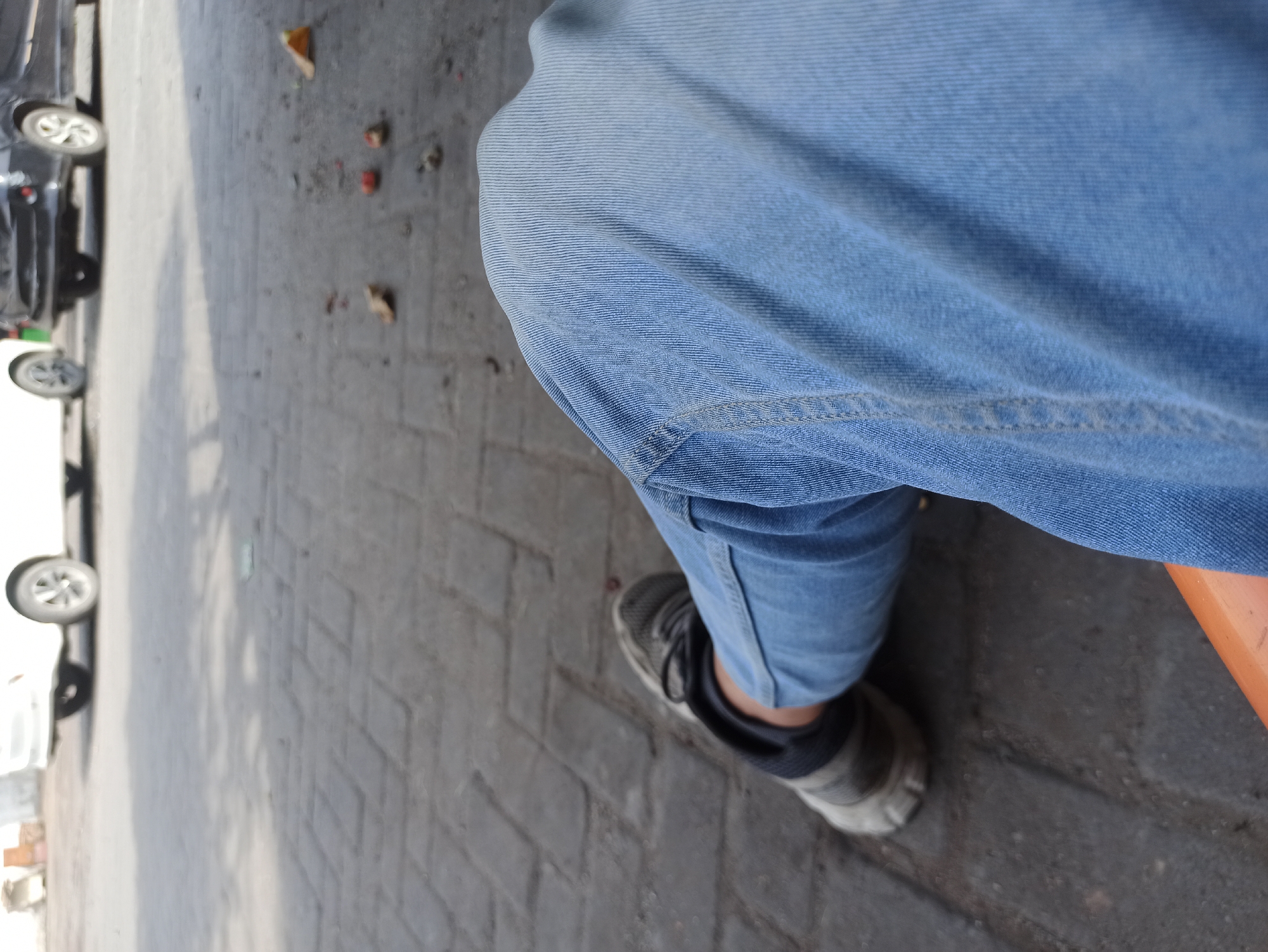 The image shows a close-up view of a person's legs and feet, with a blurred background of parked cars and a paved surface. The person is wearing light blue jeans and dark, athletic shoes with lighter gray accents. The shoes appear to be worn and a bit dirty. The background reveals the rear ends of two vehicles; a white car on the left and a dark-colored car on the right. The ground is paved with rectangular bricks or stones, and there are scattered leaves and debris visible. The lighting suggests an outdoor setting during the day, possibly on a street or parking area. The image is framed from a low angle, focusing on the lower body of the individual. There is no specific information in the image that indicates the location is the City of Medan, Indonesia.