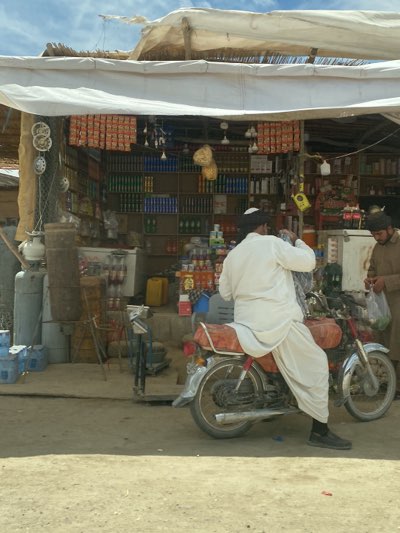 The image shows a roadside shop in Sard-E 'olya, Afghanistan. The shop, shaded by a white awning, has shelves stocked with various goods, including canned beverages, snacks, and household items. A man wearing a white shalwar kameez and a black turban is seated on a red motorcycle parked in front of the shop, appearing to be making a purchase. Another man in a brown outfit is standing next to the motorcycle, holding a bag. The area in front of the shop appears to be a dirt road, and there are metal canisters and various other objects near the shop's entrance.