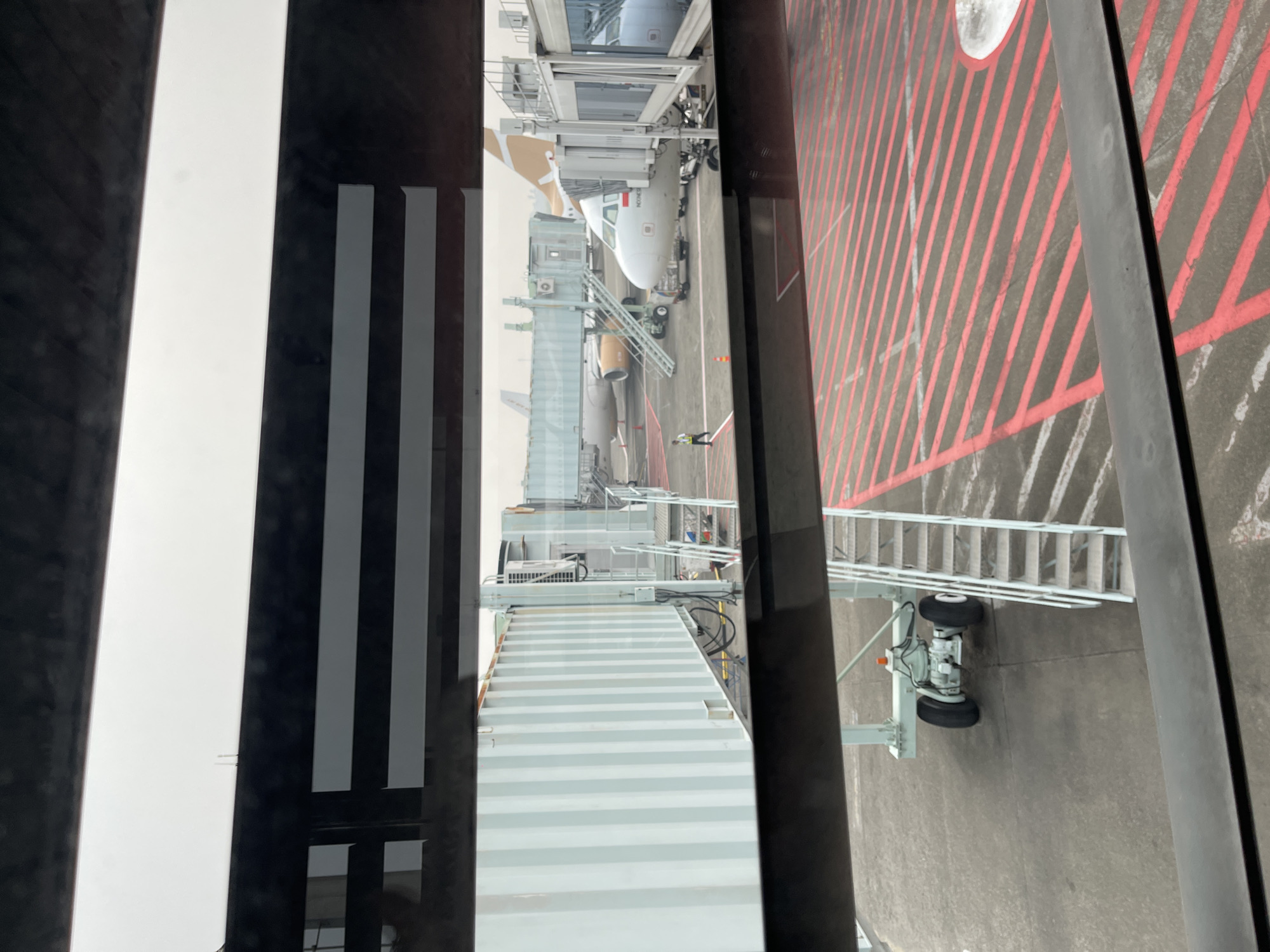 The image depicts an airport tarmac in Tangerang, Indonesia, viewed from inside a terminal building. In the foreground, a jet bridge with a light blue, ribbed exterior is connected to the terminal. Below the jet bridge, a section of the tarmac is visible with red and white striped markings, a set of stairs, and a wheeled support structure for an aircraft.
In the middle ground, several airplanes are parked at gates. One white airplane with the word "INDONESIA" and a red and white flag on its tail is connected to a jet bridge. Another airplane, predominantly white with some gold accents on its tail, is visible behind it. A single person, dressed in a dark shirt and pants, is walking on the tarmac near the jet bridge.
The background shows more of the airport infrastructure, including additional jet bridges and the hazy sky. The overall scene suggests a busy airport environment.