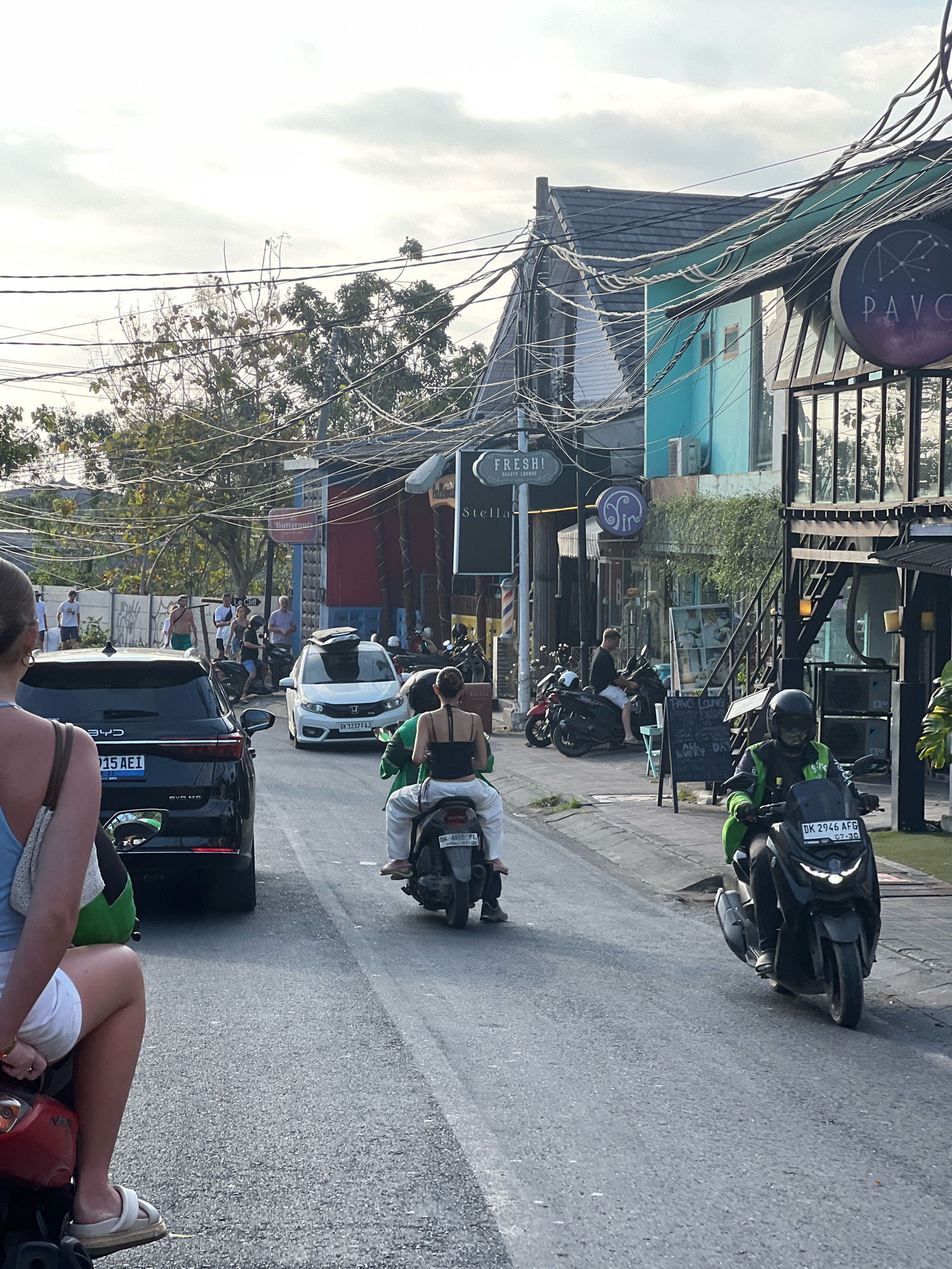 In Kuta Selatan, Indonesia, a street scene unfolds with a mix of vehicles and pedestrians. A black BYD SUV is parked on the left, facing away from the camera. In the center of the road, a white Honda Brio is moving forward. Several motorcycles are also present, with two in the foreground. The rider of the foremost motorcycle is wearing a black helmet and a green jacket, while the rider of a motorcycle further back is wearing a black helmet and a white outfit.
The street is lined with buildings displaying various signage, including "PAVO," "FRESH! BEAUTY LOUNGE," and "Stella." Electrical wires are strung across the scene, creating a dense network overhead. On the sidewalks, a few people are visible, some standing and others walking. The overall impression is of a busy urban environment with active street life. The sky is partly cloudy, suggesting it could be daytime.