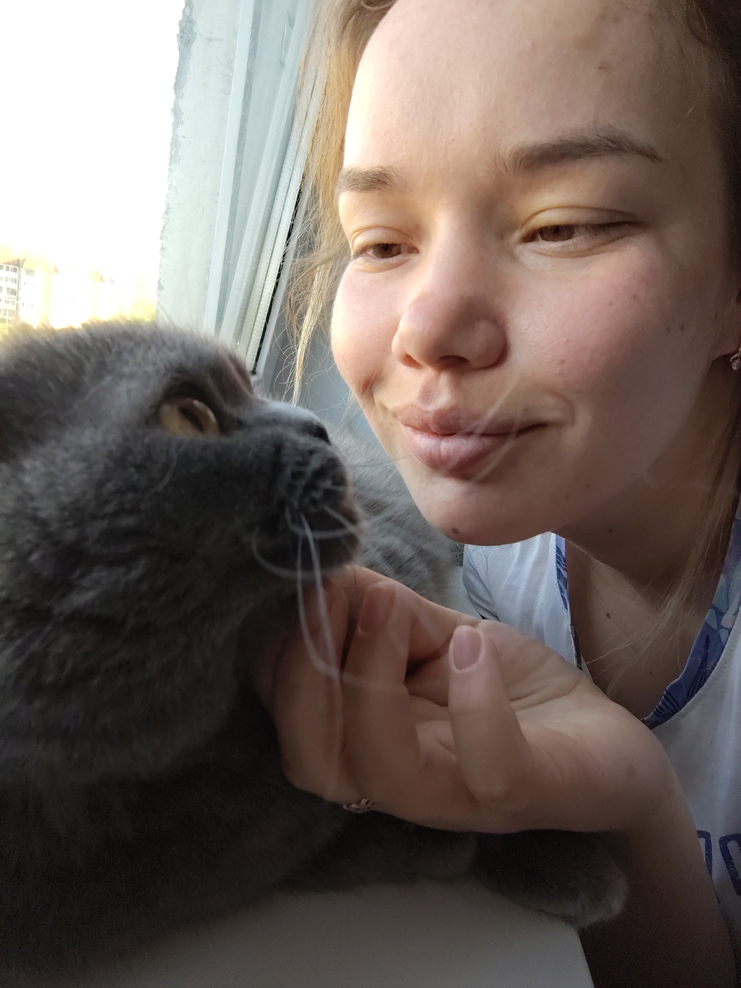 A woman with fair skin, light brown hair, and a slight smile is photographed closely with a gray cat. The woman's hand is touching the cat's face, near its whiskers. They are indoors, near a window with buildings visible outside. The photo was taken in the city of Марковский, Russia.