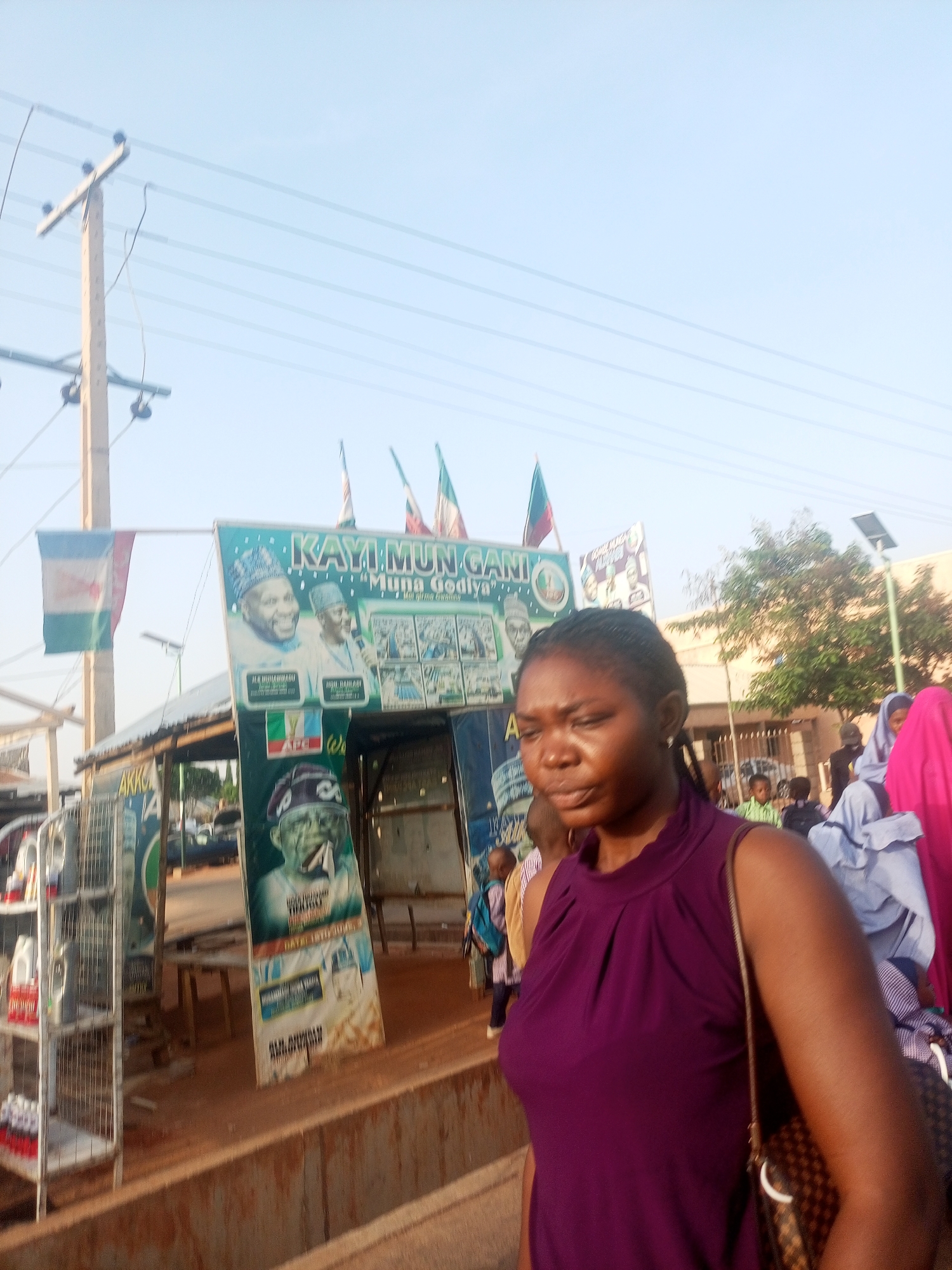 The image depicts a street scene in an urban location. In the foreground, a dark-skinned woman wearing a purple sleeveless top and carrying a patterned handbag is partially visible.
Behind her, there is a makeshift structure or kiosk decorated with political posters and flags. One prominent sign reads "KAYI:MUN GANI" and displays images of political figures. The letters "APC" are also visible on the sign. Other people, including some wearing headscarves, are milling about in the background.
Further back, there's a building and some trees. A utility pole with power lines extends into the sky on the left side of the frame. The time of day appears to be late afternoon or early evening, based on the soft lighting.