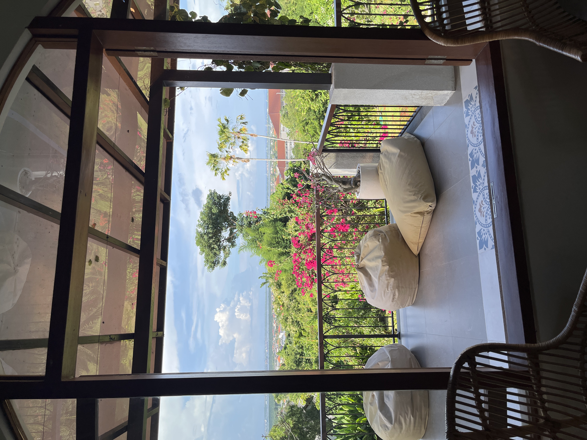From an elevated vantage point in Kuta Selatan, Indonesia, the image shows a partially covered balcony space with three large, light-colored beanbag chairs arranged on gray tiled flooring. A black wrought-iron railing adorned with vibrant pink bougainvillea separates the balcony from a lush, green landscape, featuring trees and vegetation leading down to a distant ocean view under a blue, partly cloudy sky. Two slender palm trees stand out in the mid-ground. The balcony is framed by dark wooden beams and the interior of a room, where the texture of rattan chairs are visible. The view encompasses the urban sprawl of Kuta Selatan.