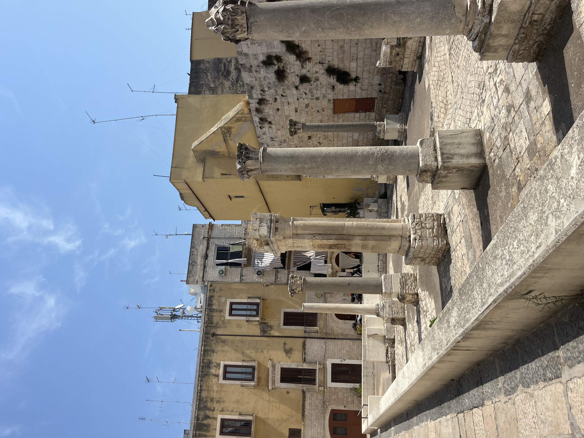 In the city of Bari, Italy, the image displays an outdoor archaeological site featuring ancient Roman columns. The columns, some damaged and incomplete, stand on a cobblestone ground surrounded by historic buildings. The buildings have a mixture of rough stone and painted plaster facades, with wooden doors and windows, and balconies. The sky above is blue with scattered clouds. Cell phone towers and antennas are visible on the rooftops of the buildings, indicating a blend of ancient and modern elements within the urban landscape. There are no people or animals present in the scene.