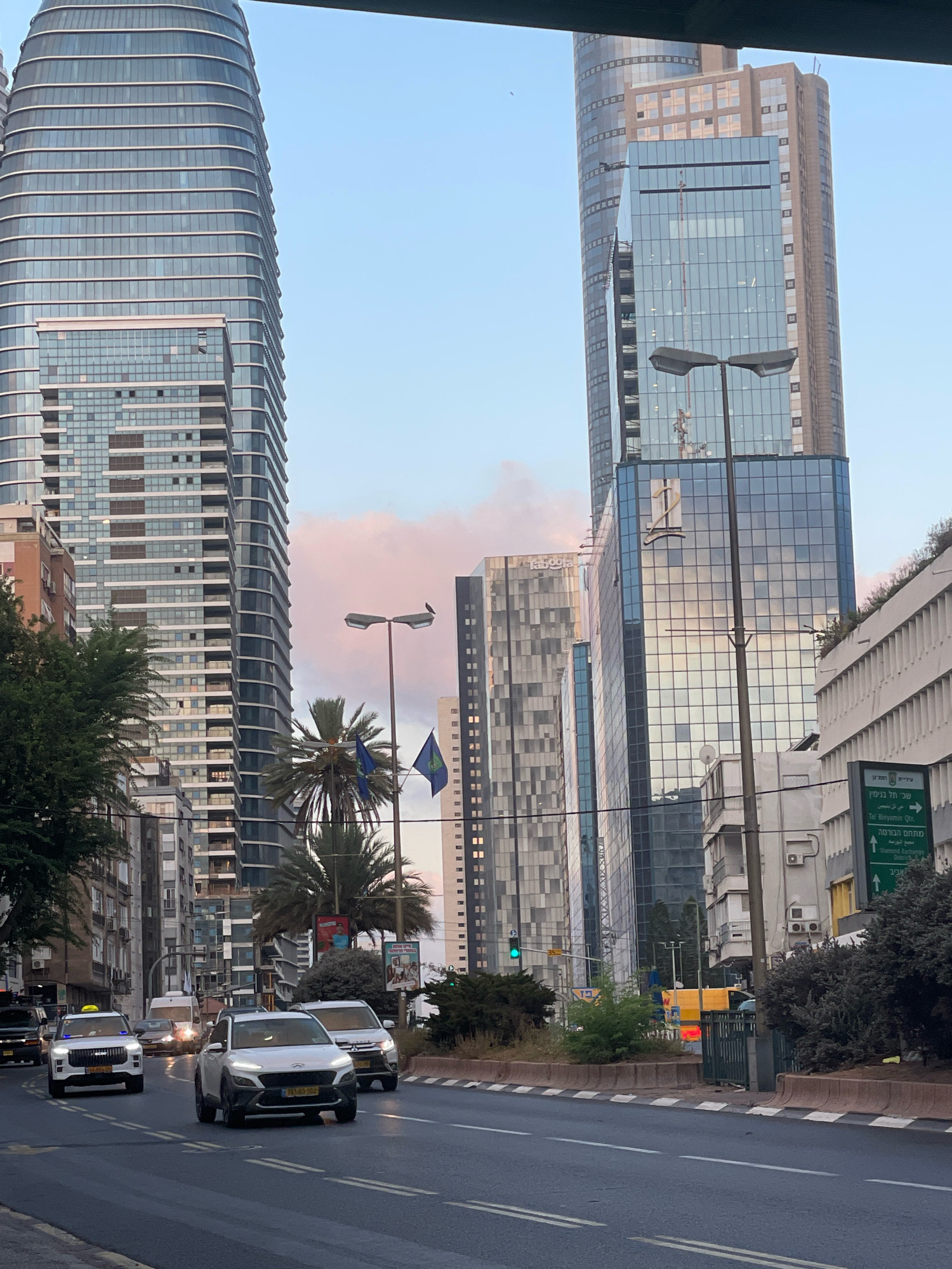 The image depicts a street scene in Ramat Gan, Israel, characterized by modern architecture and traffic. In the foreground, a multi-lane road displays cars in motion, with a white SUV prominently positioned in the center lane. The road surface appears wet, suggesting recent rain or cleaning. On the left side of the road, a white taxi with blue flashing lights is visible, and behind it, other vehicles are also present.
The backdrop is dominated by tall, contemporary buildings. A distinctive, curved skyscraper with a glass facade stands on the left, its many floors reflecting the sky. To its right, another taller skyscraper with a mix of glass and opaque sections rises, featuring a large "12" logo on its facade. Other buildings of varying heights and architectural styles are also part of the urban landscape, some with visible windows and balconies.
Vegetation, including palm trees and bushes, is interspersed throughout the scene, particularly along the median of the road and on sidewalks. Streetlights and traffic signals are also visible, indicating a functional urban infrastructure. The sky exhibits a soft, pastel hue, with hints of pink and blue, suggesting it might be either dawn or dusk. A few flags, one blue and one green, are attached to a lamppost, adding a touch of color to the scene. There are no people clearly visible in the image, but the presence of cars and buildings implies ongoing urban activity.