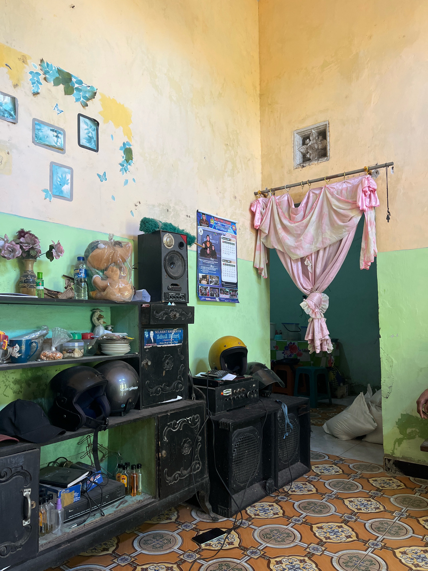 The image depicts an interior room with a distinct cultural aesthetic, likely in a residential setting in Ngablak, Indonesia. The walls are painted in a muted yellow and vibrant green, showing signs of age and wear. Decorative elements include faded blue floral decals and butterflies on the yellow wall, and a small, square ornate carving above a draped pink curtained doorway.
The room is filled with a variety of objects. A green shelf unit displays several items, including a vase of artificial flowers, bottles, a teddy bear in plastic, a stack of plates, and several jars and containers. Below this, a dark, ornate wooden cabinet houses two motorcycle helmets, a black baseball cap, various bottles of liquids, and electronic equipment including a DVD player. Adjacent to the cabinet are two large black speaker cabinets, one topped with a yellow helmet and electronic controls. A calendar is affixed to the wall to the right of the speaker.
The floor is covered in patterned tiles featuring warm orange and brown tones with intricate floral motifs in blue and white. In the background, through the draped doorway, a glimpse of another room reveals a teal wall, a green plastic stool, and several large white sacks stacked on the floor. The lighting suggests it is daytime, casting natural light into the room. No people are clearly visible within the frame, though a hand is partially visible on the right edge of the image. The overall impression is one of a lived-in, eclectic space, reflecting local Indonesian décor and lifestyle.