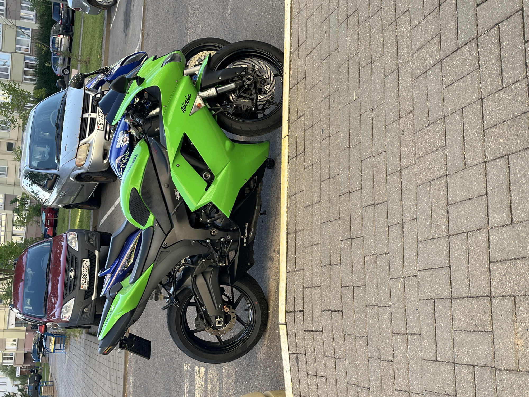 Two motorcycles are parked side-by-side on a paved surface in a city. The motorcycle in the foreground is bright green with "Ninja" written on the fairing. The second motorcycle, partially obscured by the first, appears to be blue and black. Behind the motorcycles, a red Lada car with Belarusian license plate "5488 CC-7" is parked, and a silver minivan with a Belarusian license plate "B174 K" is also visible. The background shows apartment buildings and trees, suggesting an urban residential area. The pavement is made of interlocking grey bricks. The scene appears to be outdoors during daylight.