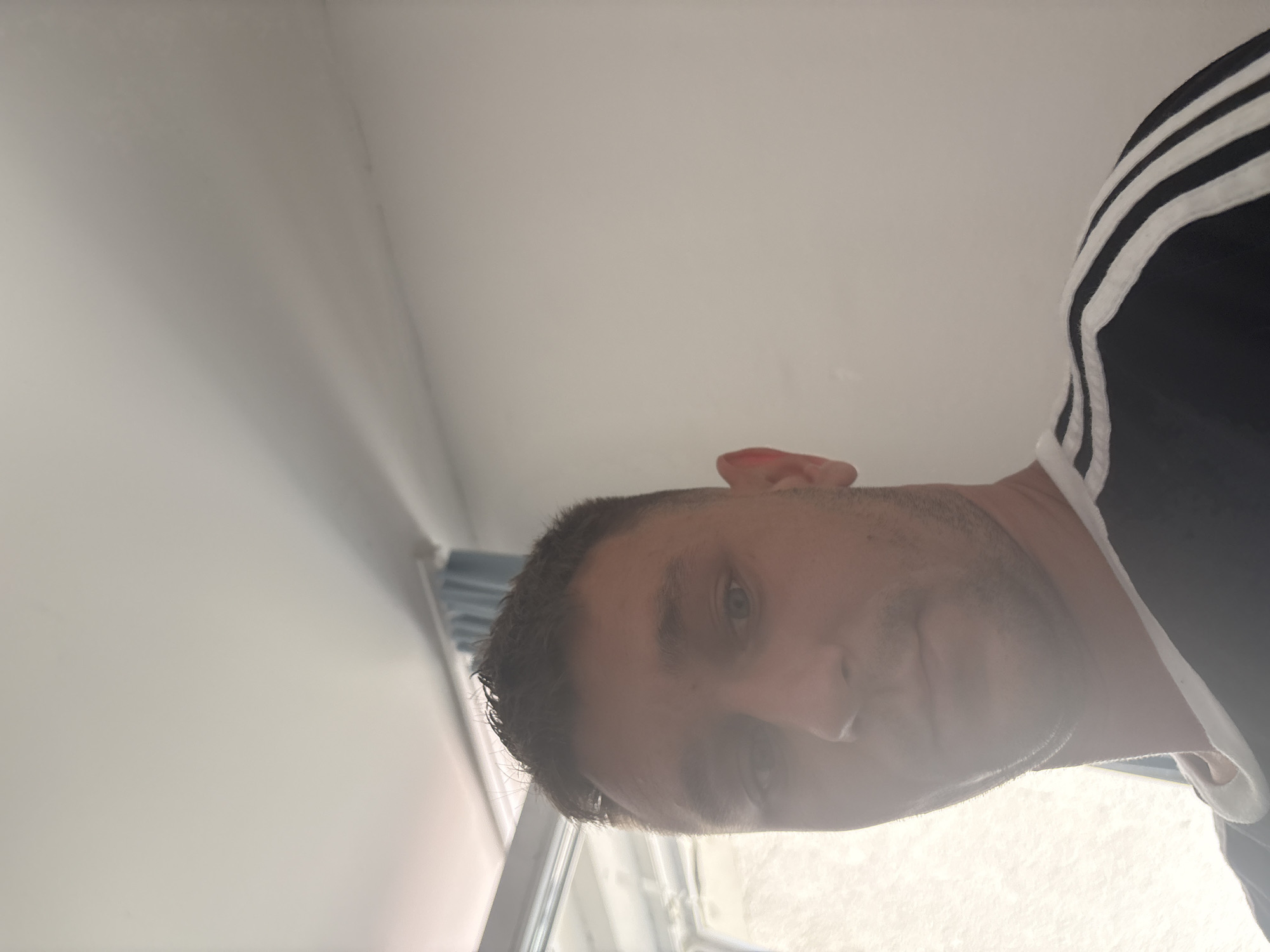 This media file is a portrait of a person from a low-angle perspective, as if looking up at them. The subject is a fair-skinned individual with short, dark hair, and they are wearing a black shirt with white stripes on the shoulder. The person's face is partially obscured by a bright light source, but their eyes, nose, and mouth are visible. They appear to be looking directly into the camera with a neutral expression. The background is a plain, light-colored wall, with a window or doorway visible on the left side of the frame. The lighting suggests it is daytime, possibly indoors near a window. There is no visible text in the image.