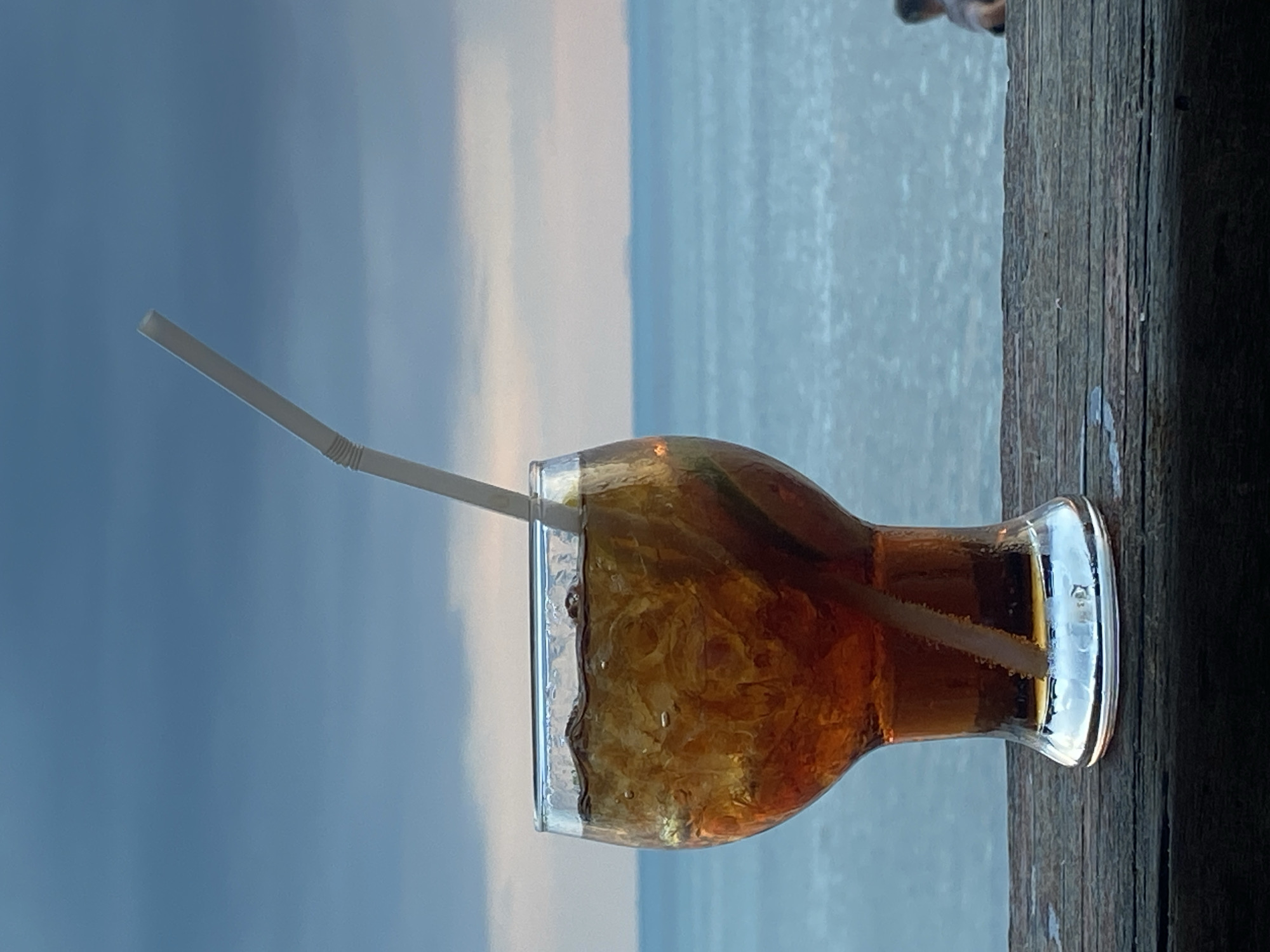 A glass of cola with ice and a straw sits on a wooden surface. In the background, the ocean meets a cloudy sky. The location is Ko Samui District, Thailand. A person is partially visible in the far right background.