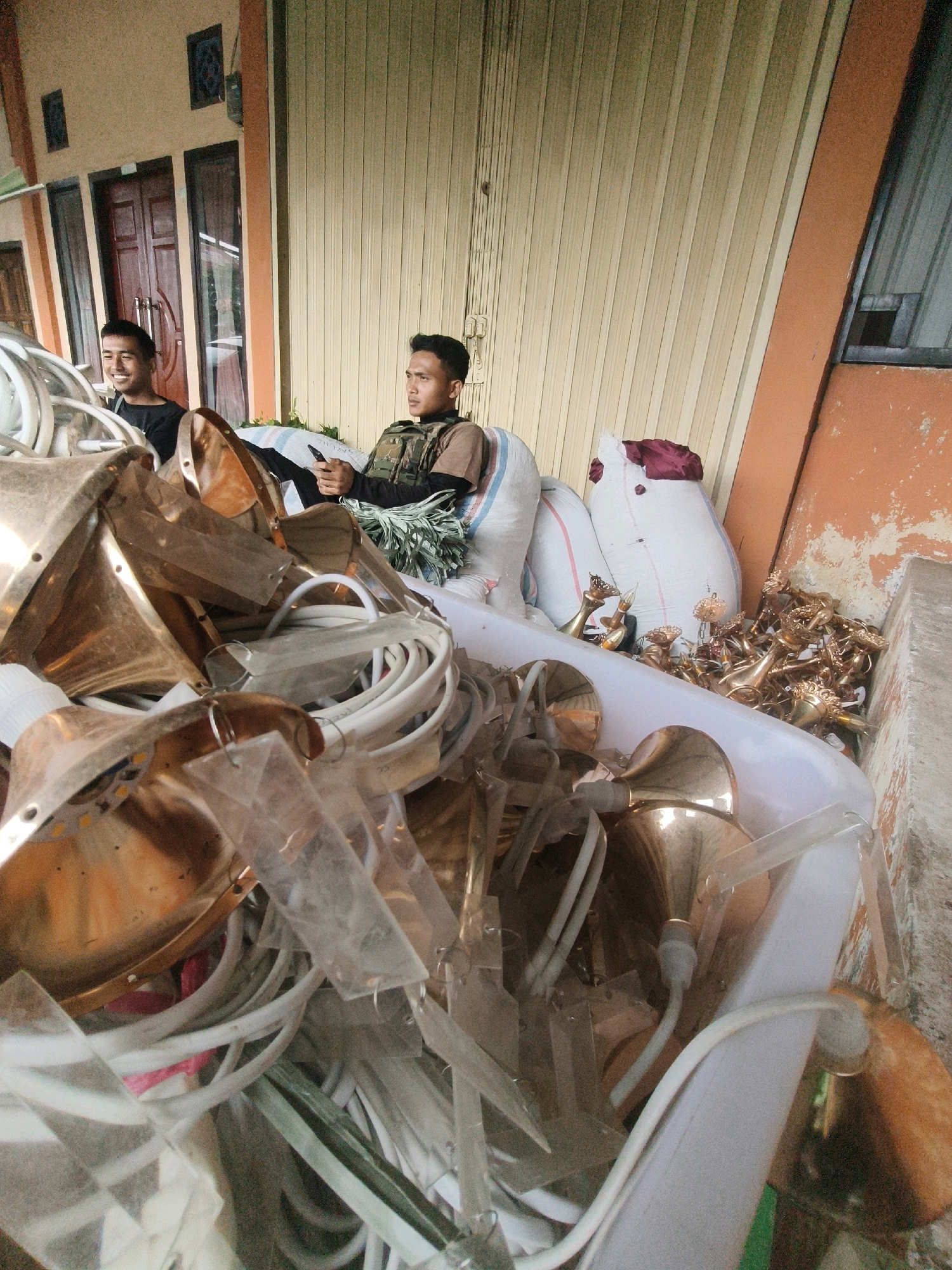 The image captures a scene outside a building with an orange and beige facade in Lindung Jaya, Indonesia. In the foreground, a large white plastic container is filled with a jumbled assortment of metallic objects, primarily brass-colored lampshades and associated electrical wiring. Scattered amongst these are also white LED bulbs and clear plastic protective coverings.
Behind the container, two men are seated. The man on the left is wearing a black t-shirt and appears to be smiling. The man on the right is wearing a tactical vest over a brown shirt and is holding a mobile phone, looking to his left. They are seated on large sacks, one white with red stripes and another white, with a dark red cloth draped over it.
The background features a building with a corrugated metal door, windows, and what appear to be wooden doors. The overall environment suggests an outdoor storage or workshop area.