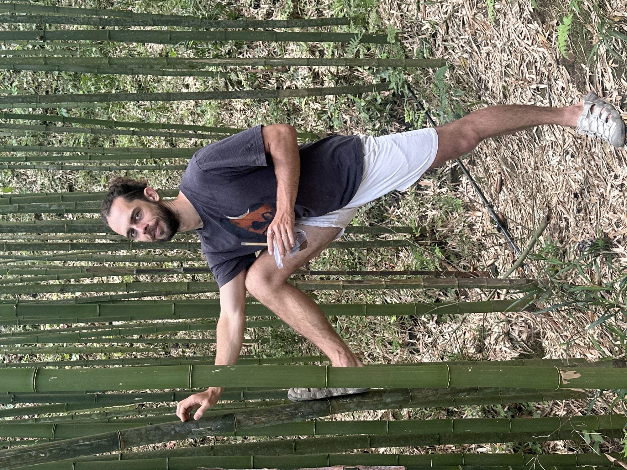 The image shows a man standing in a bamboo forest in Ngã Ba Kim, Vietnam. He is wearing a dark t-shirt, white shorts, and one grey shoe. He appears to be holding a small clear container and touching a bamboo stalk with his left hand. The ground is covered in dry leaves and vegetation. Bamboo stalks surround him.