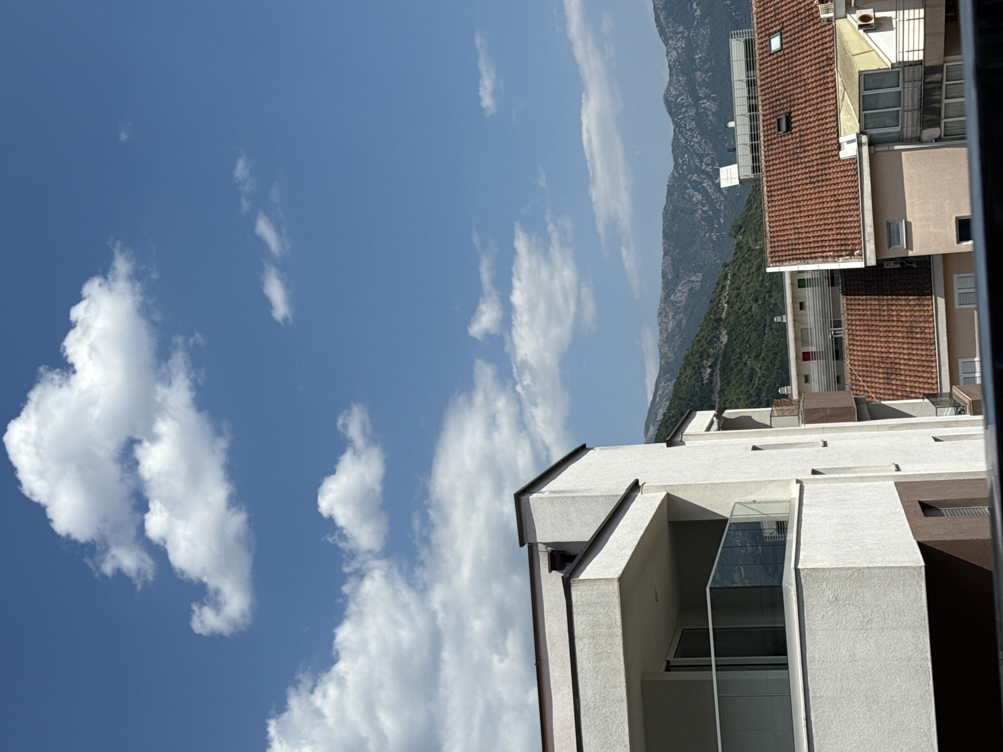 The image captures a daytime view in Budva, Montenegro, looking out over a residential area towards mountains. In the foreground, the side of a modern, off-white building with a brown base and a balcony featuring a glass railing is prominently displayed. Adjacent to it, other buildings with terracotta tiled roofs are visible. The background is dominated by lush green, tree-covered mountains under a bright blue sky with scattered white clouds. The overall scene depicts a sunny day in a coastal city with mountainous terrain.